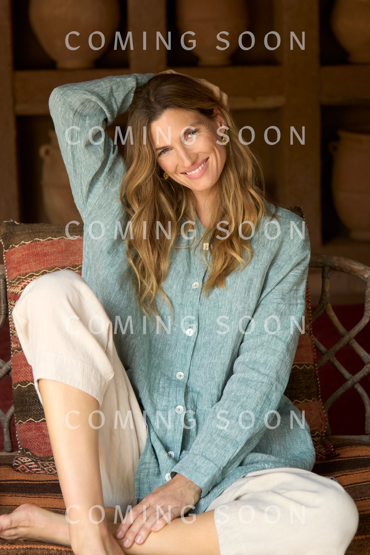 A woman with wavy hair, wearing a light green button-up shirt and Twisted Linen Crop Bubble Trouser, sits on a cushioned bench and smiles at the camera. The words "COMING SOON" overlay the image.