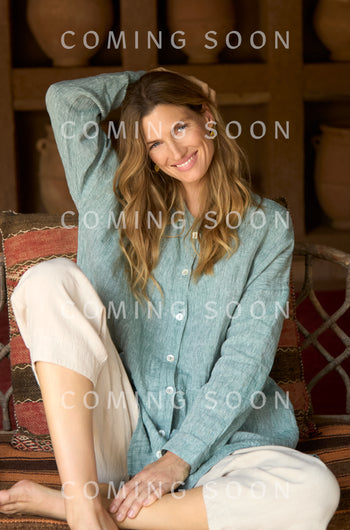 A woman with wavy hair, wearing a light green button-up shirt and Twisted Linen Crop Bubble Trouser, sits on a cushioned bench and smiles at the camera. The words "COMING SOON" overlay the image.