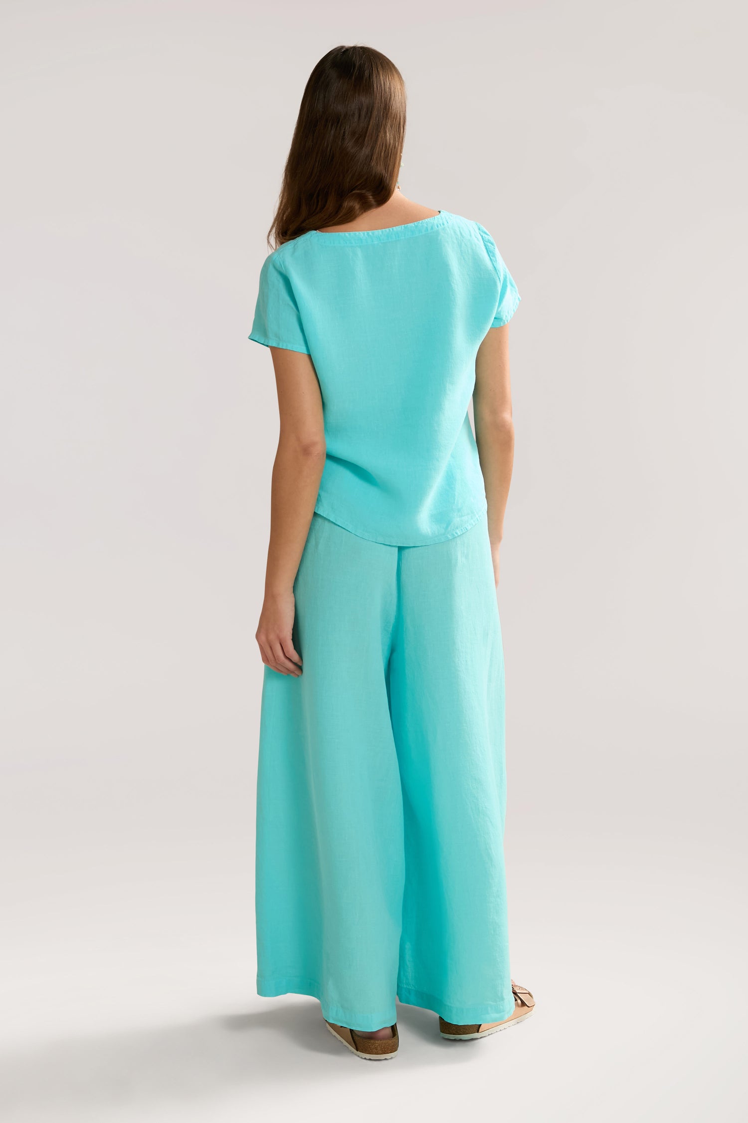 A woman with long hair, seen from the back, is wearing a light blue short-sleeve top and matching Organza Linen Wide Trouser that offer a touch of relaxed luxury with their elasticated waist.