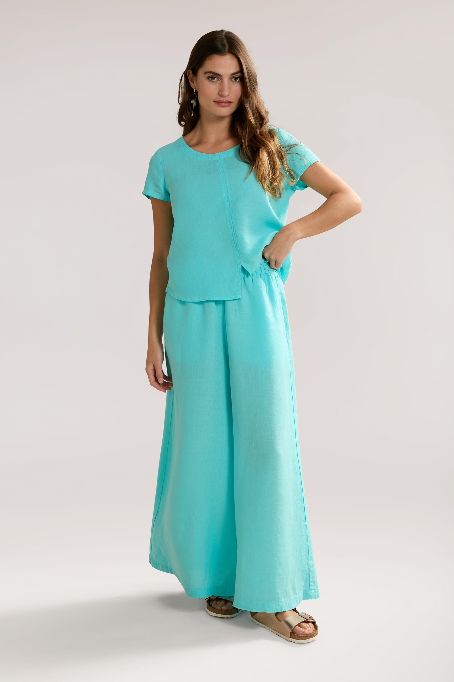 A woman wearing a turquoise two-piece outfit with Organza Linen Wide Trousers featuring an elasticated waist and short sleeves stands against a plain background, effortlessly embodying relaxed luxury.