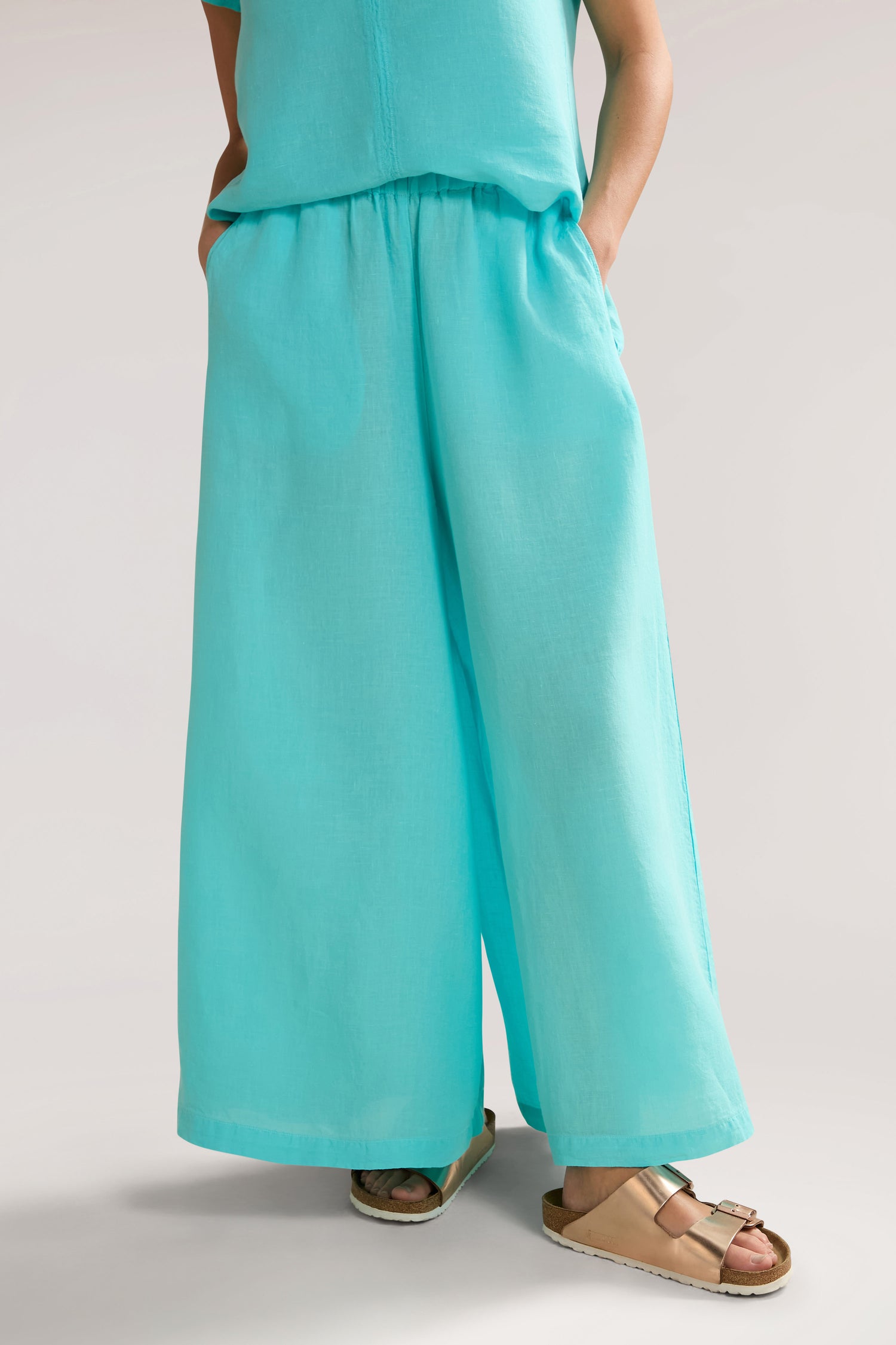 Person wearing turquoise Organza Linen Wide Trouser and beige sandals stands with hands in pockets, exuding a relaxed luxury.