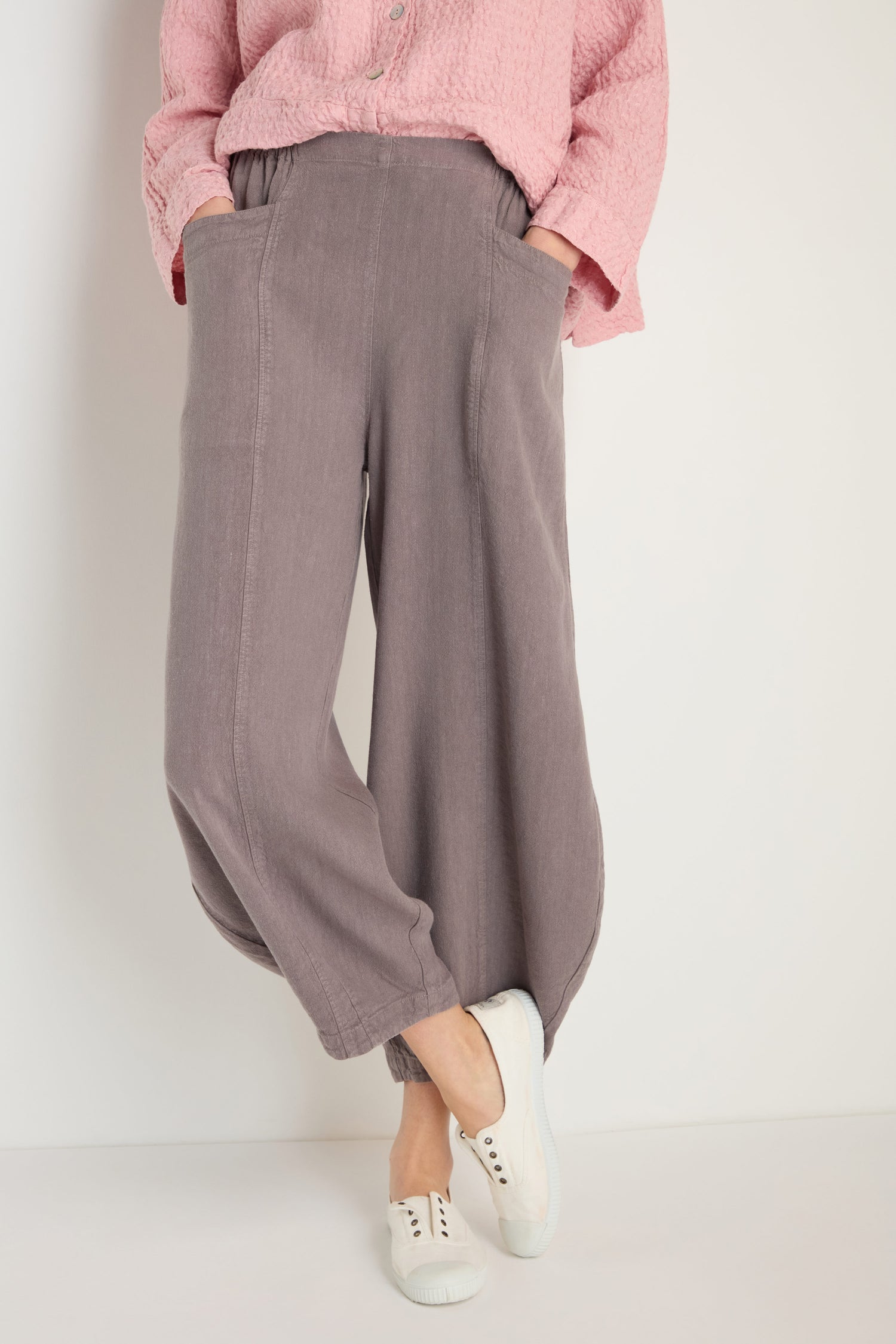 Person wearing Twisted Linen Bubble Trouser in breathable grayish-purple with elastic cuffs, paired with a textured pink top and white slip-on shoes, standing against a plain background.