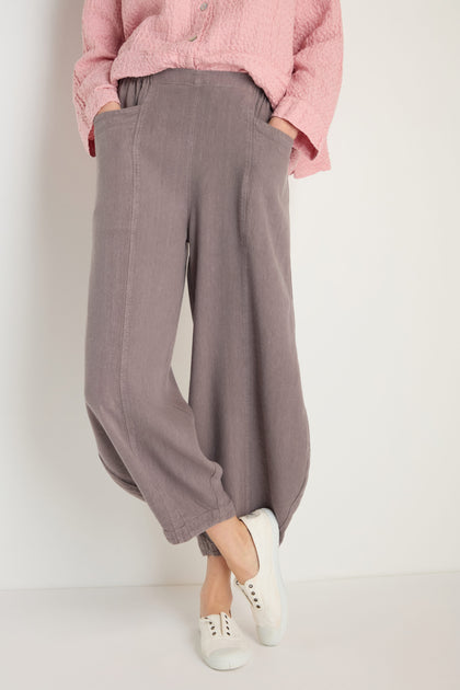 Person wearing Twisted Linen Bubble Trouser in breathable grayish-purple with elastic cuffs, paired with a textured pink top and white slip-on shoes, standing against a plain background.