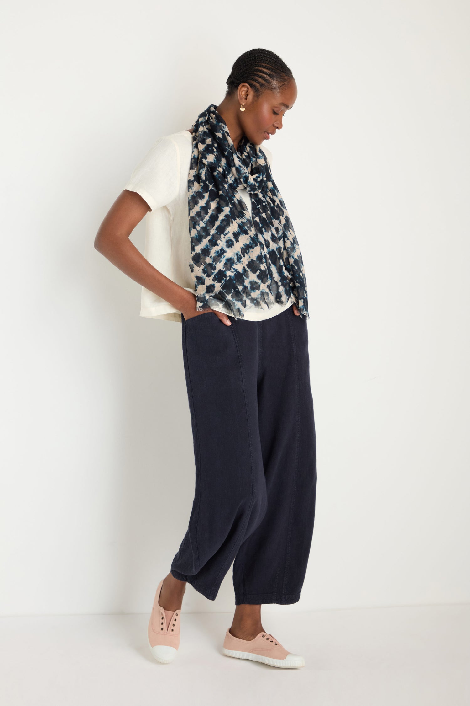 A woman stands against a white wall wearing a cream breathable short-sleeve top, Twisted Linen Bubble Trousers in dark color, a patterned scarf, and pink slip-on shoes.