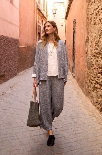 A woman wearing loose grey Crinkle Double Face Linen Bubble Trousers, a jacket, white top, and black shoes carries a tote bag as she walks down a narrow sunlit alley with pink walls.