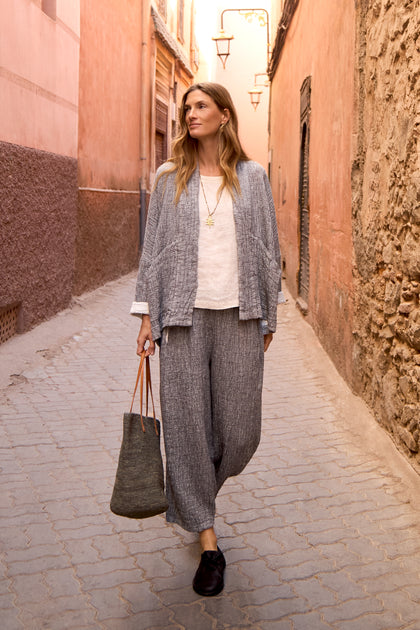 A woman wearing loose grey Crinkle Double Face Linen Bubble Trousers, a jacket, white top, and black shoes carries a tote bag as she walks down a narrow sunlit alley with pink walls.
