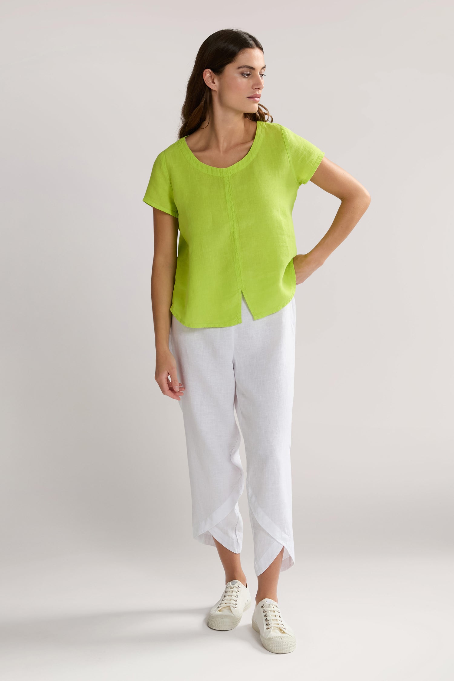 A woman in a lime green Slit Hem Linen Top and white pants stands with one hand on her hip, looking to the side. She is wearing white sneakers.