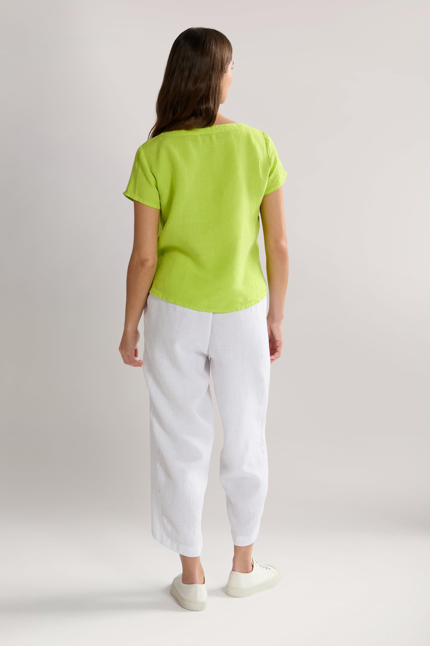 Woman standing and facing away, wearing a Slit Hem Linen Top in lime green with flutter sleeves, white pants, and white shoes, with a plain light-colored background.