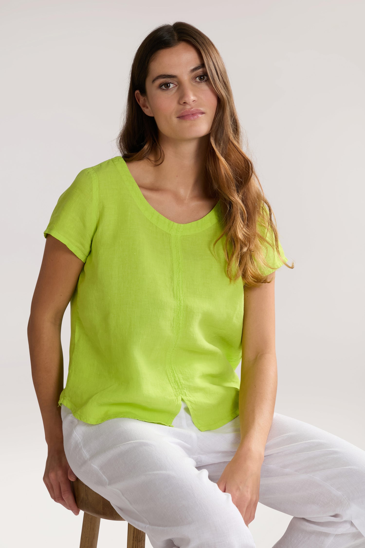 A woman with long brown hair wearing a bright green short-sleeved Slit Hem Linen Top and white pants sits on a wooden stool against a plain background.