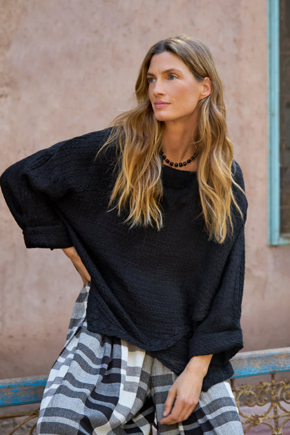 A woman with long, wavy hair wears the Waffle Linen Boxy Cowl Neck Top in black with checkered pants, sitting by a textured pale wall and a blue-framed window.