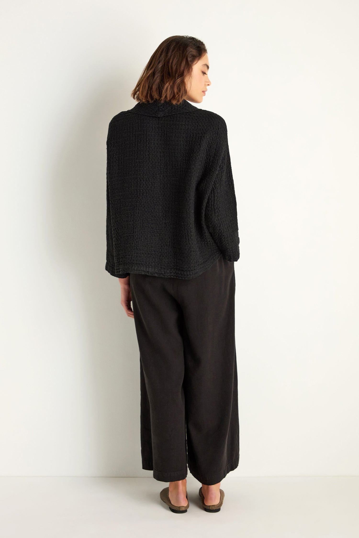 A woman faces away from the camera wearing the Waffle Linen Boxy Cowl Neck Top, styled with wide-leg black pants and flat shoes, against a plain white background.