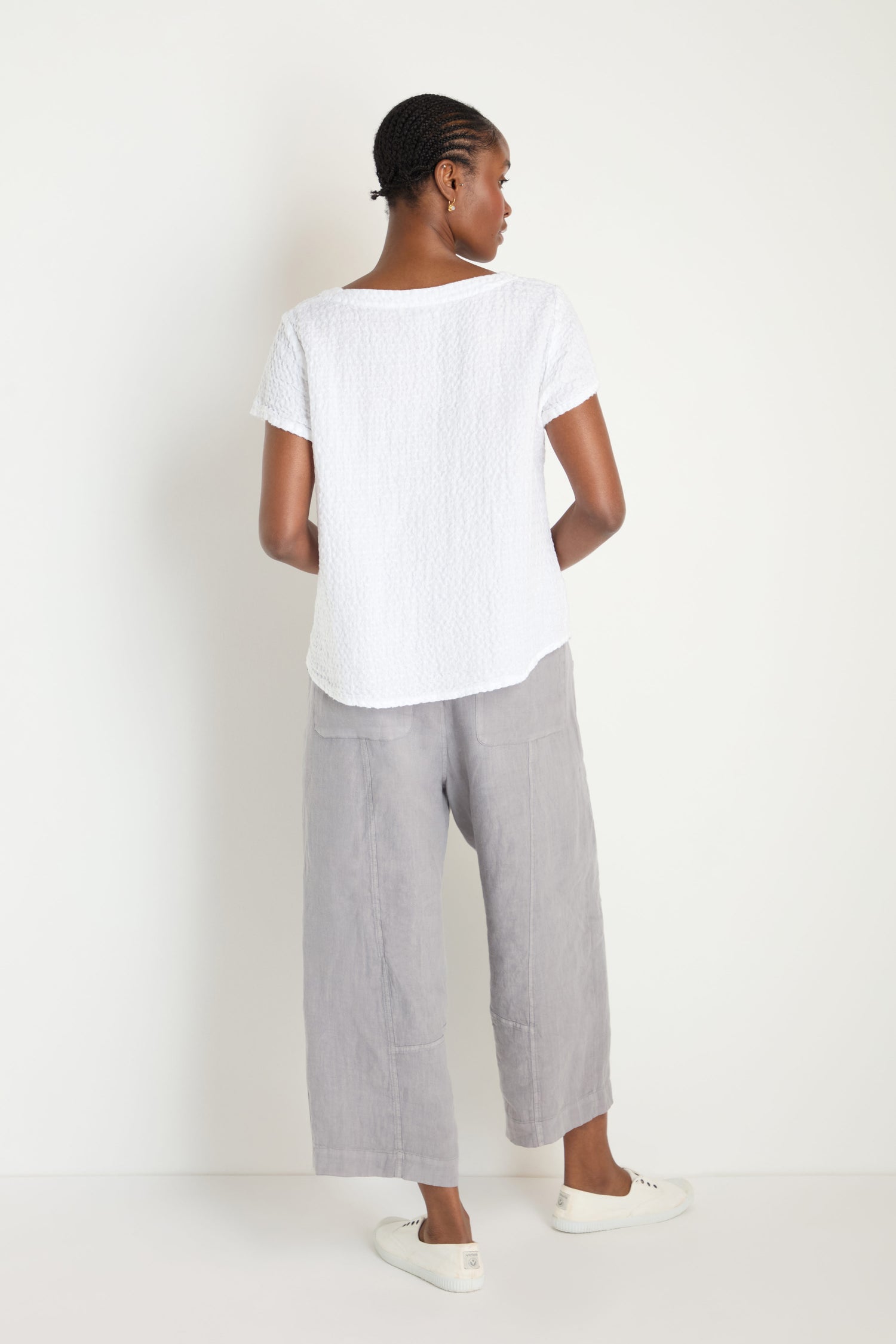 A person stands facing away, wearing a white short-sleeve Waffle Linen V Neck Top, light gray wide-leg pants, and white slip-on shoes against a plain white background.
