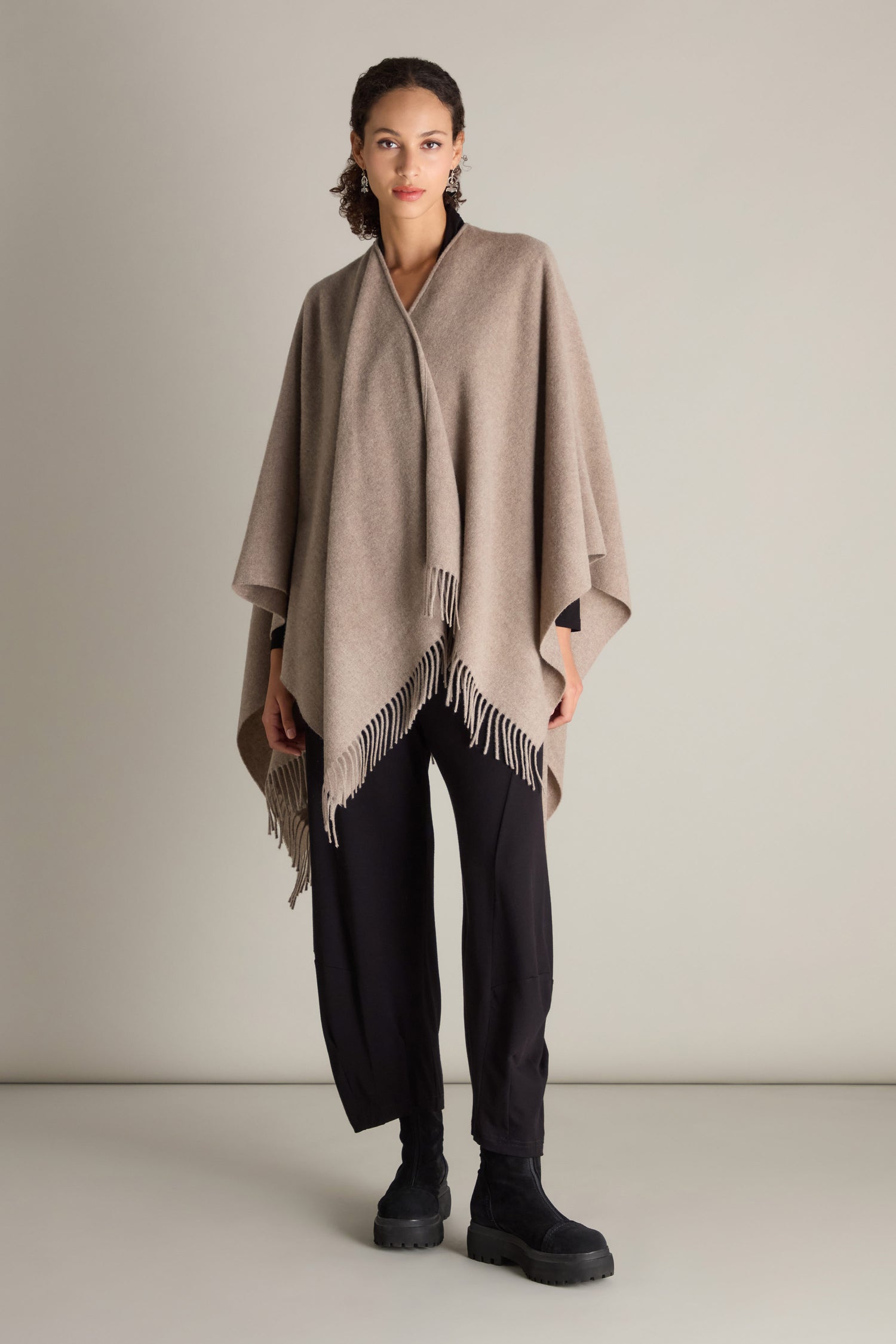 A person stands wearing the Wool Cashmere Poncho with fringe detailing, paired with black pants against a plain background.