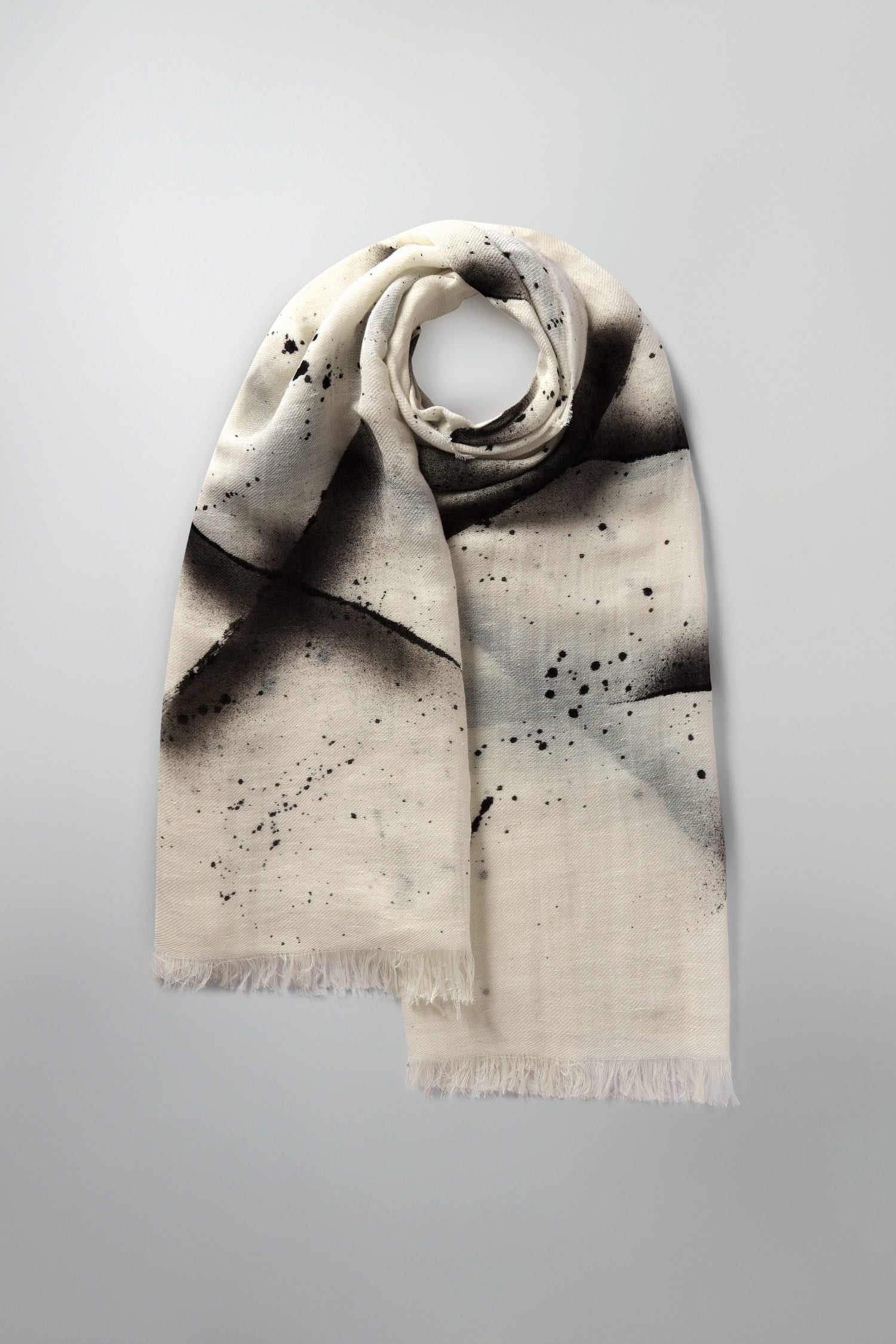 The Abstract Brushstroke Scarf is a perfect spring wardrobe addition, showcasing a striking abstract black splatter and brushstroke pattern on a gray background. It is elegantly crafted from a luxurious linen and cotton blend.