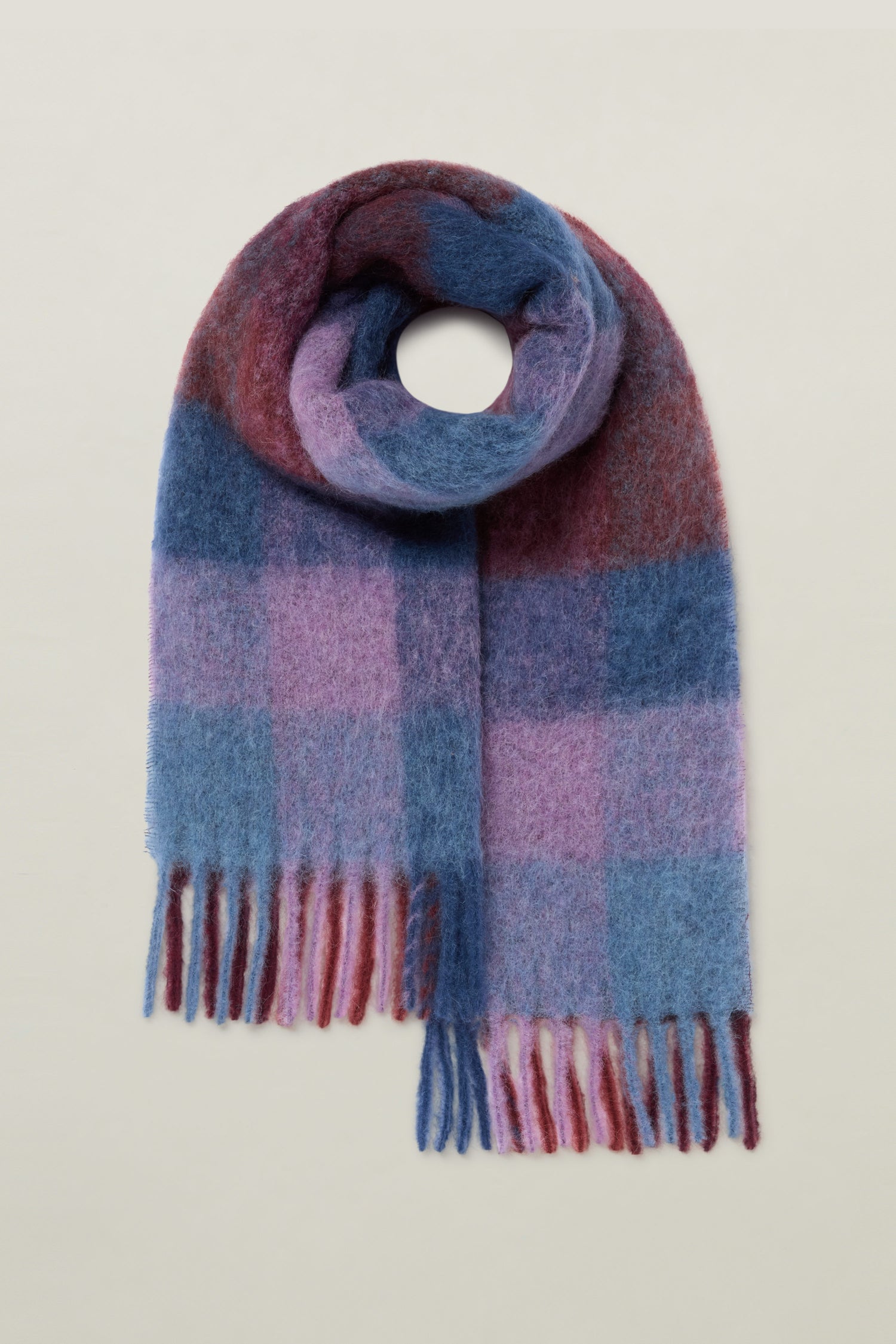 The Check Tassel Scarf, designed in a classic check pattern, is crafted from a cozy mohair wool blend. It features shades of blue, purple, and red, elegantly arranged in a loop shape with fringed ends for the perfect touch.