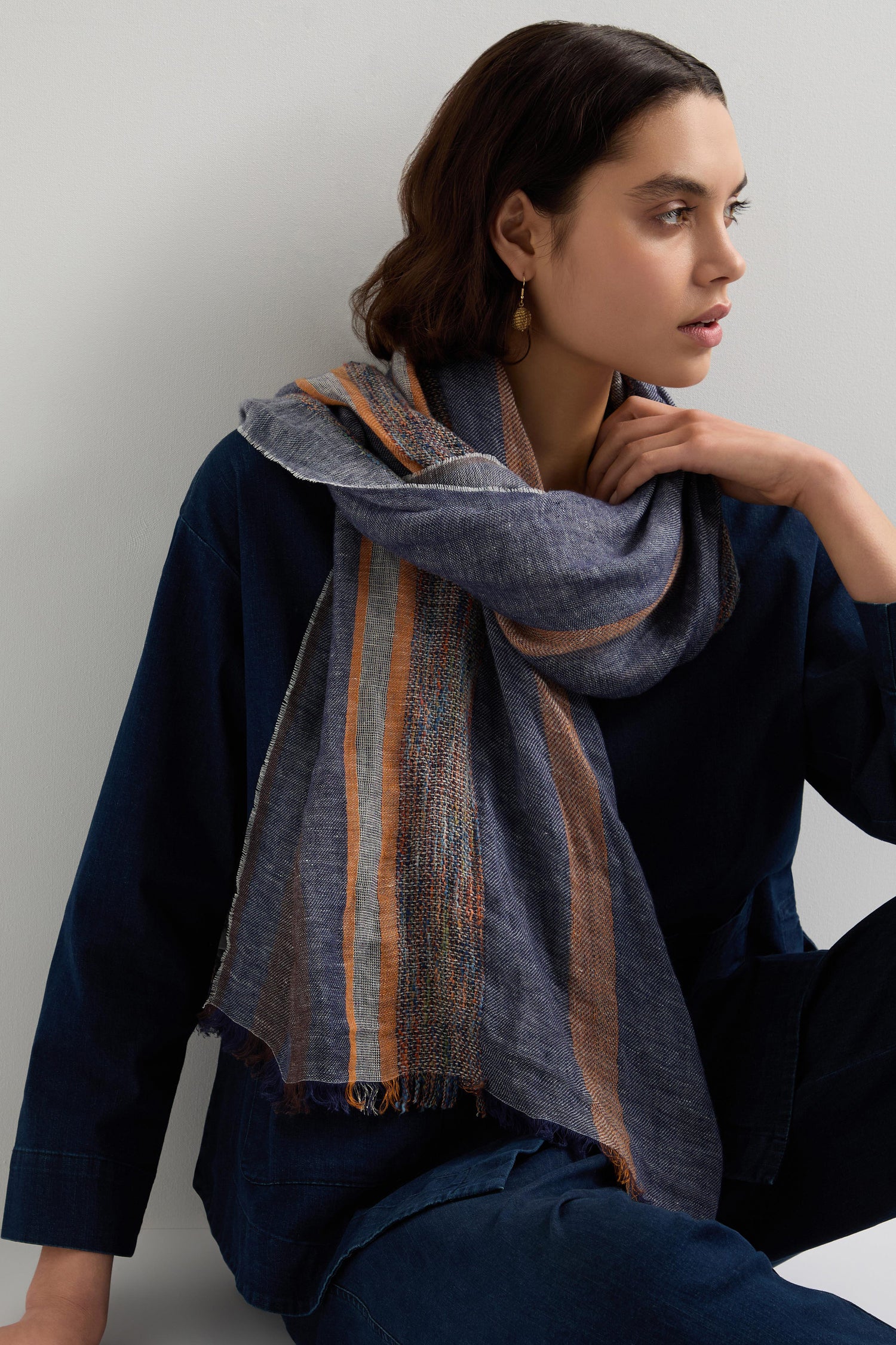 Wearing a blue outfit and a stylish Draped Scarf with orange accents and Denim Detail Linen Stripe, they sit gazing thoughtfully to the side. The linen-cotton blend ensemble perfectly enhances their chic spring and summer wardrobe with effortless elegance.