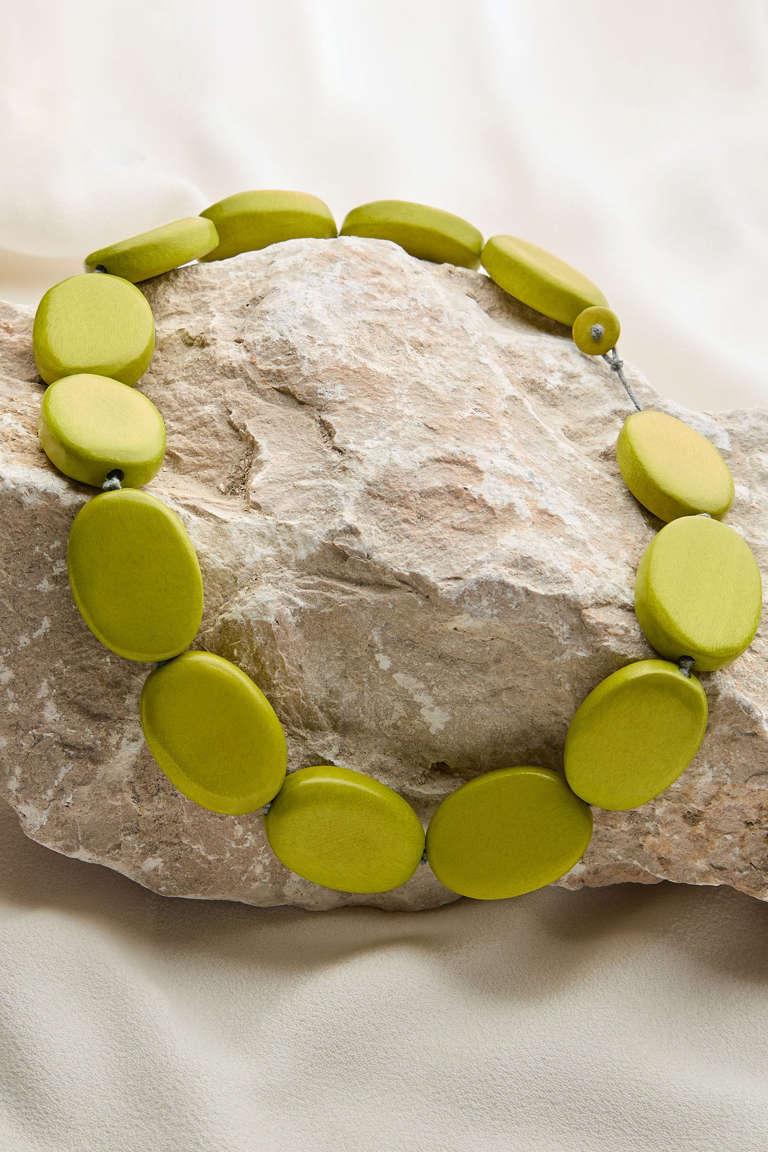 A Short Wooden Pebble Necklace, marked with the code LFH2113, is arranged on a large, textured rock with a beige, draped fabric in the background.