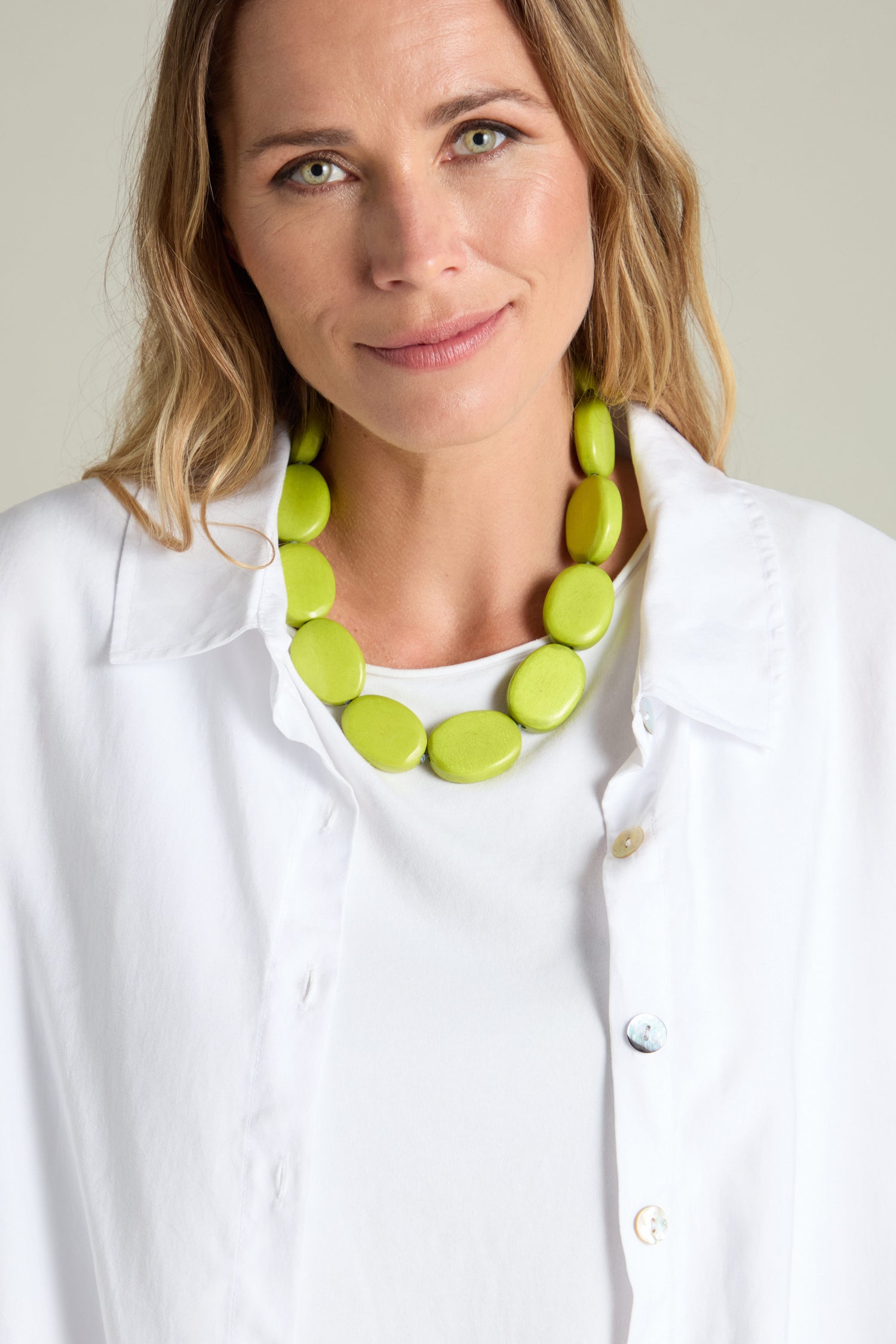 A woman with light-colored hair wears a white shirt and a Short Wooden Pebble Necklace featuring chunky wood beads, looking at the camera with a neutral expression.