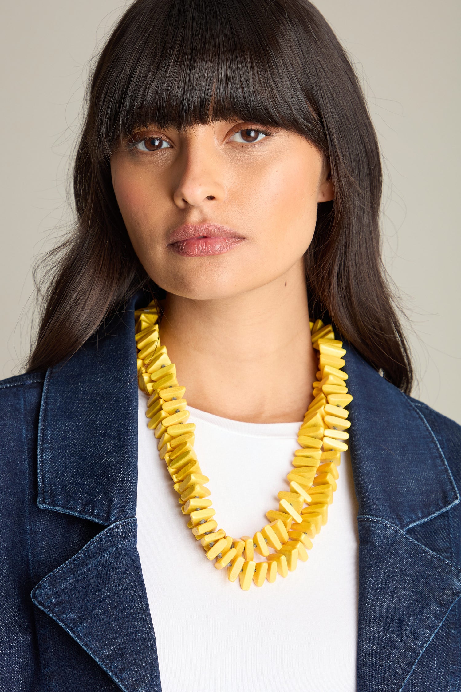 A woman wearing a white shirt, a blue jacket, and the Mini Triangles Wooden Necklace is looking at the camera with a neutral expression. Her Lotusfeet sandals peek out from under her jeans.