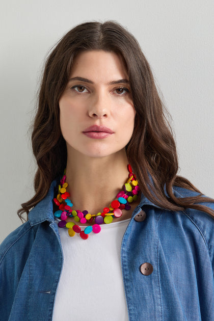 A woman with long hair, dressed in a blue denim shirt and white top, showcases the vibrant Rainbow Coconut Cascade Necklace against a plain background.