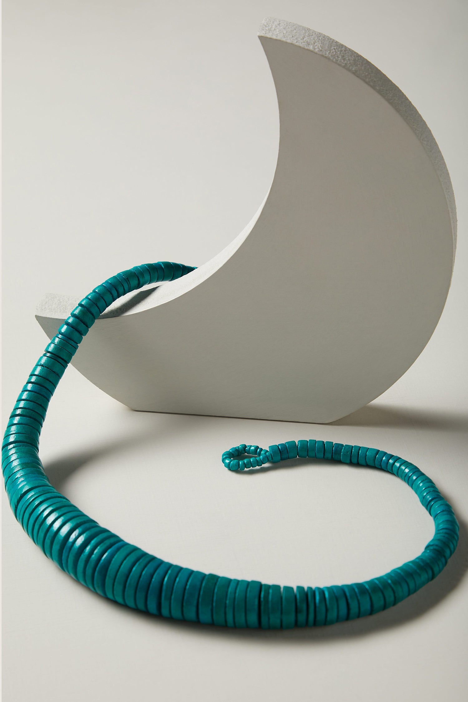 A Graduated Coco Disc Necklace, in a bold hue of turquoise, is coiled around a crescent-shaped white stand on a light-colored surface.