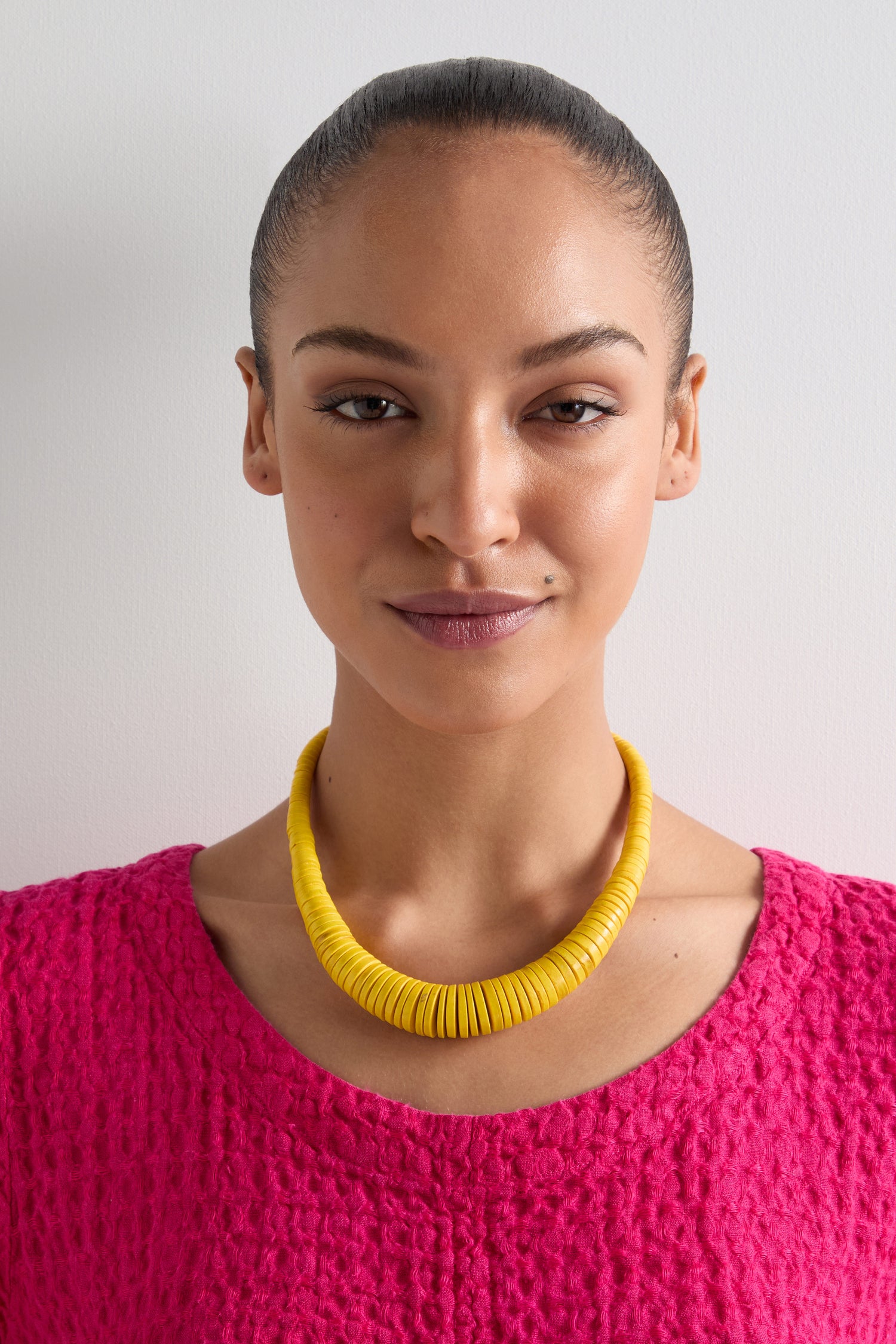 A person with a neutral expression wears a vibrant pink textured top and a Graduated Coco Disc Necklace, ethically crafted with Filipino artistry, standing against a plain white background.