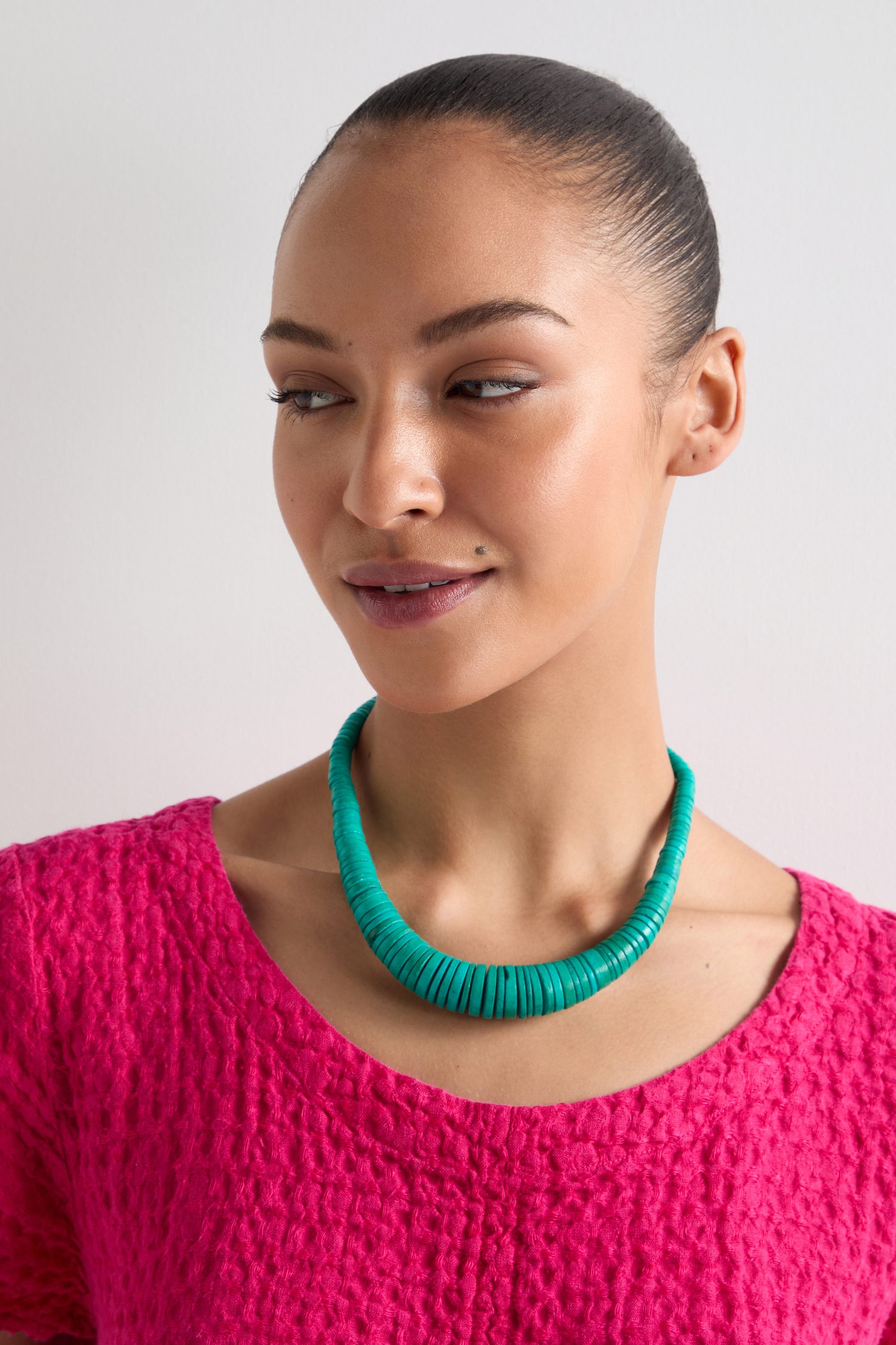 A person wearing a bright pink textured top and the Graduated Coco Disc Necklace, ethically sourced and showcasing exquisite craftsmanship from the Philippines, gazes slightly to the side against a plain background.