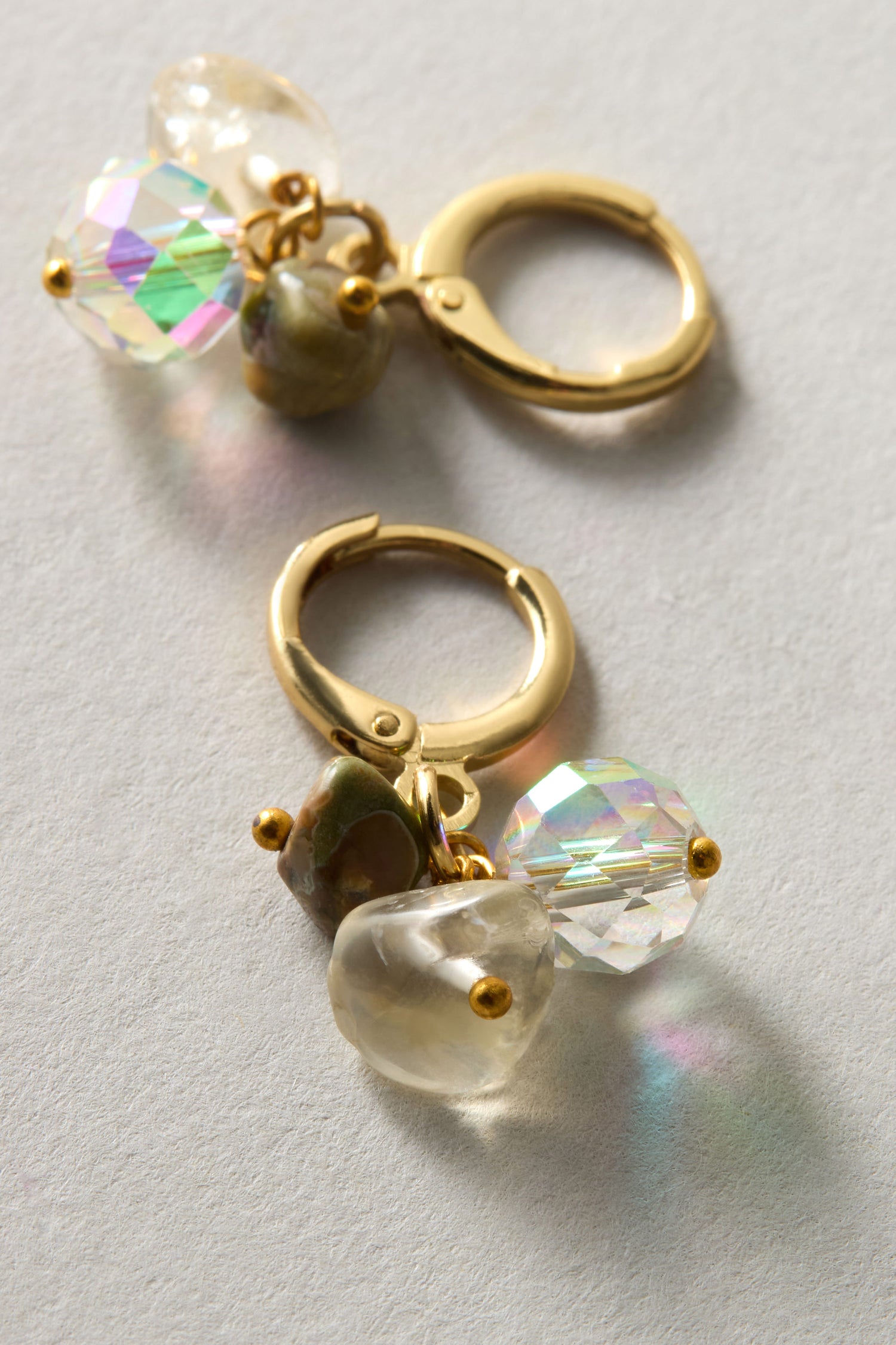 The Citrine and Rhyolite Bead Earrings feature gold hoops with iridescent rhyolite beads and polished stone-like charms, exemplifying sustainable craftsmanship on a textured surface.