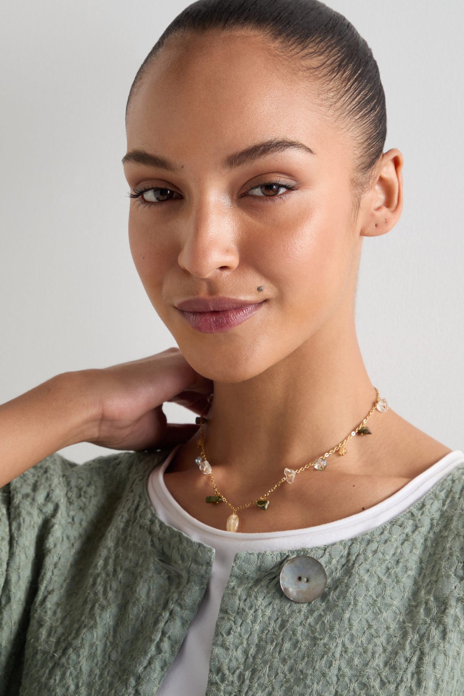 Wearing a green jacket and white top, a person dons the Citrine and Rhyolite Bead Necklace, smiling softly with one hand near their neck.