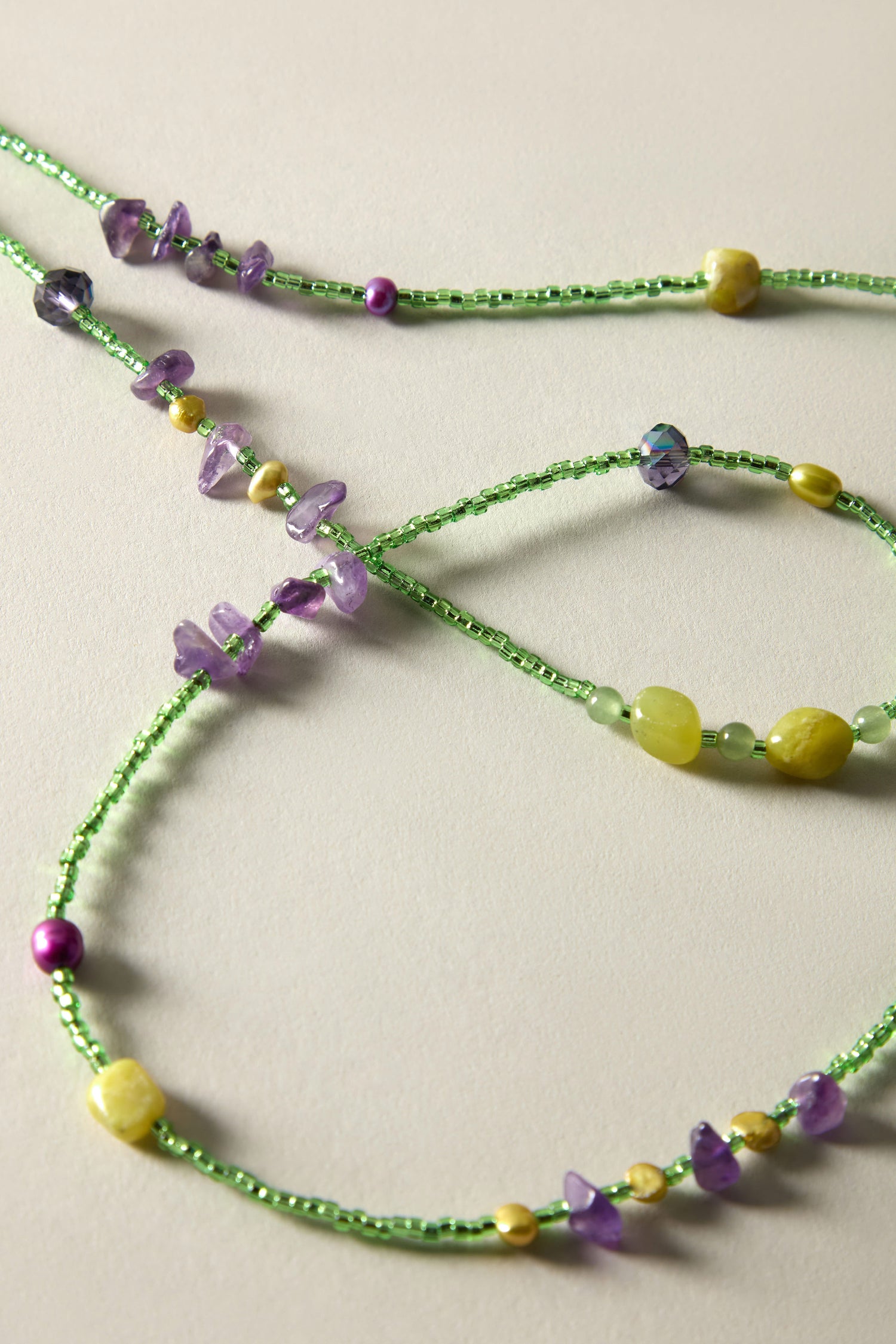 The Long Bead Necklace, with delicate green beads and interspersed purple and yellow stones, is intricately arranged on a beige surface for a bohemian-inspired statement.