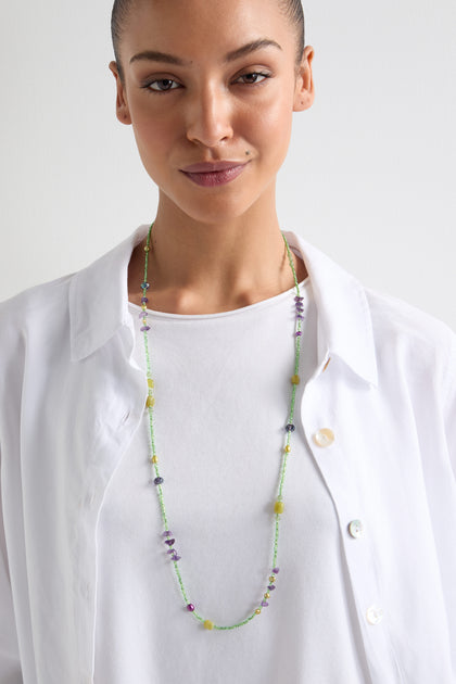 A person in a white shirt is wearing the Long Bead Necklace, featuring handcrafted green and purple beads, against a plain background. This bohemian-inspired piece adds elegance and uniqueness to their look.