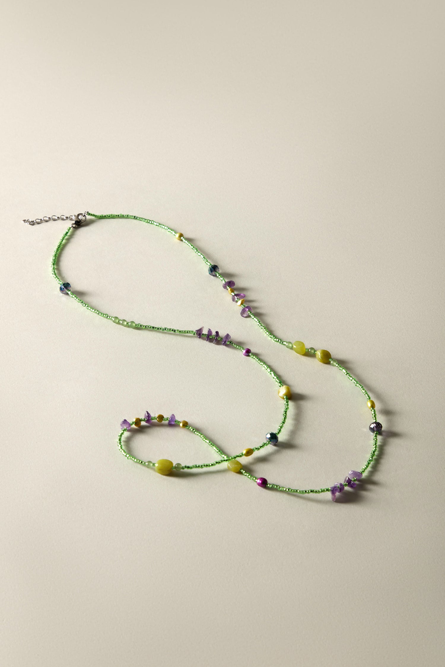 The Long Bead Necklace is a handcrafted, bohemian-inspired jewelry piece featuring artfully arranged green beads with scattered yellow, purple, and dark accents on a beige surface.