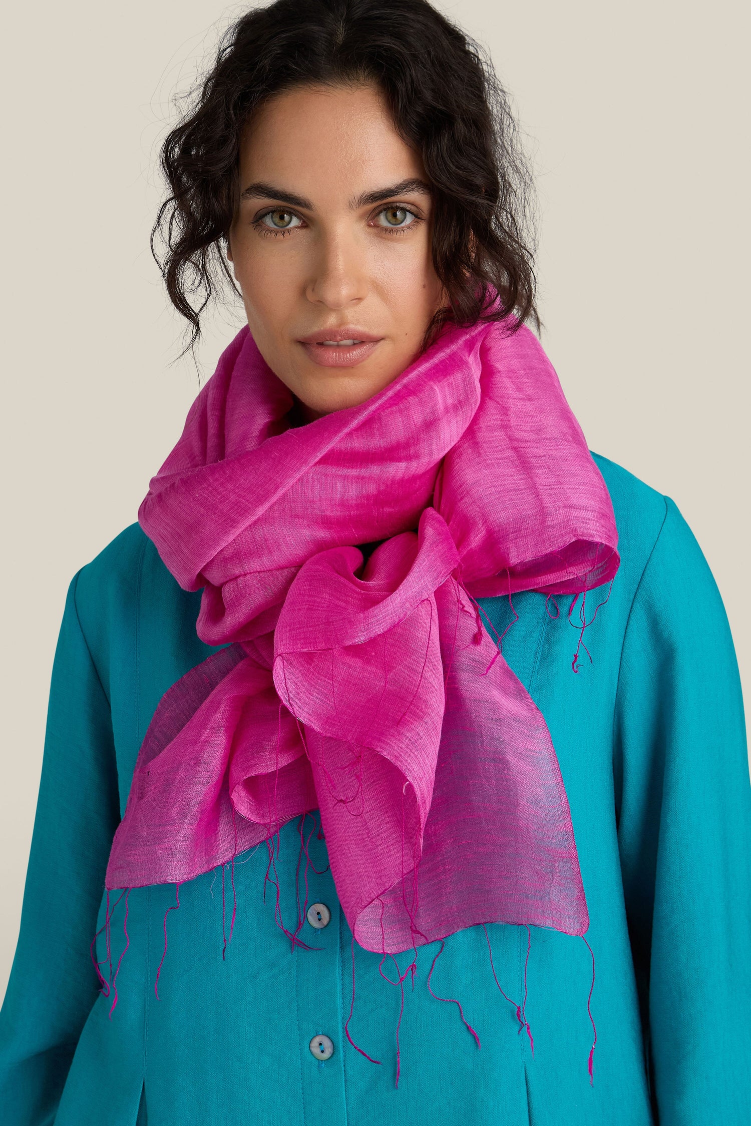 A woman with curly hair wears a bright, fair trade Soft Linen Scarf in pink over a teal top, standing against a neutral background.