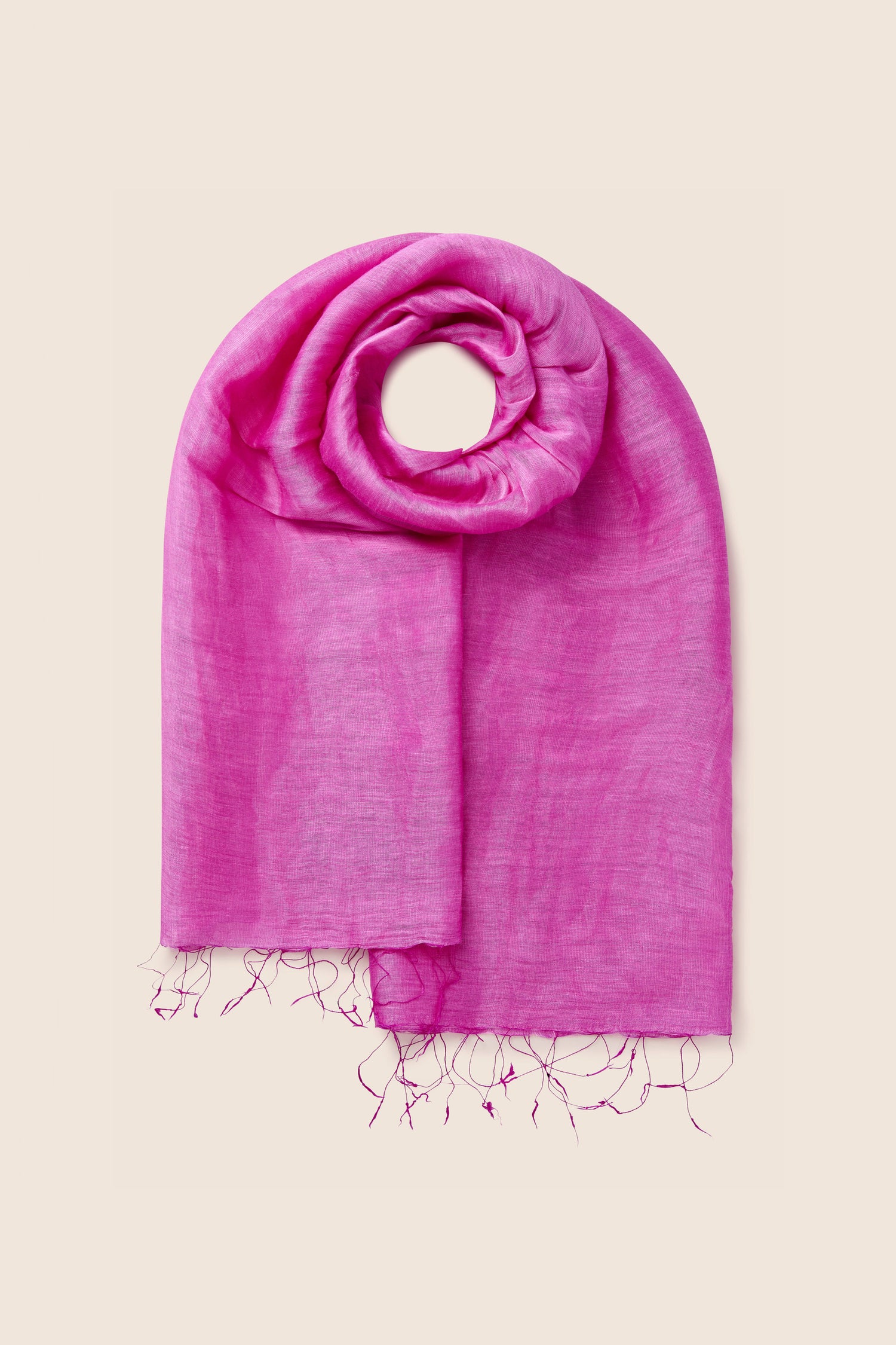 The Soft Linen Scarf, a luxurious bright pink accessory crafted from soft linen and featuring frayed ends, is elegantly arranged in a circular fashion on a beige background to highlight its fair trade craftsmanship.