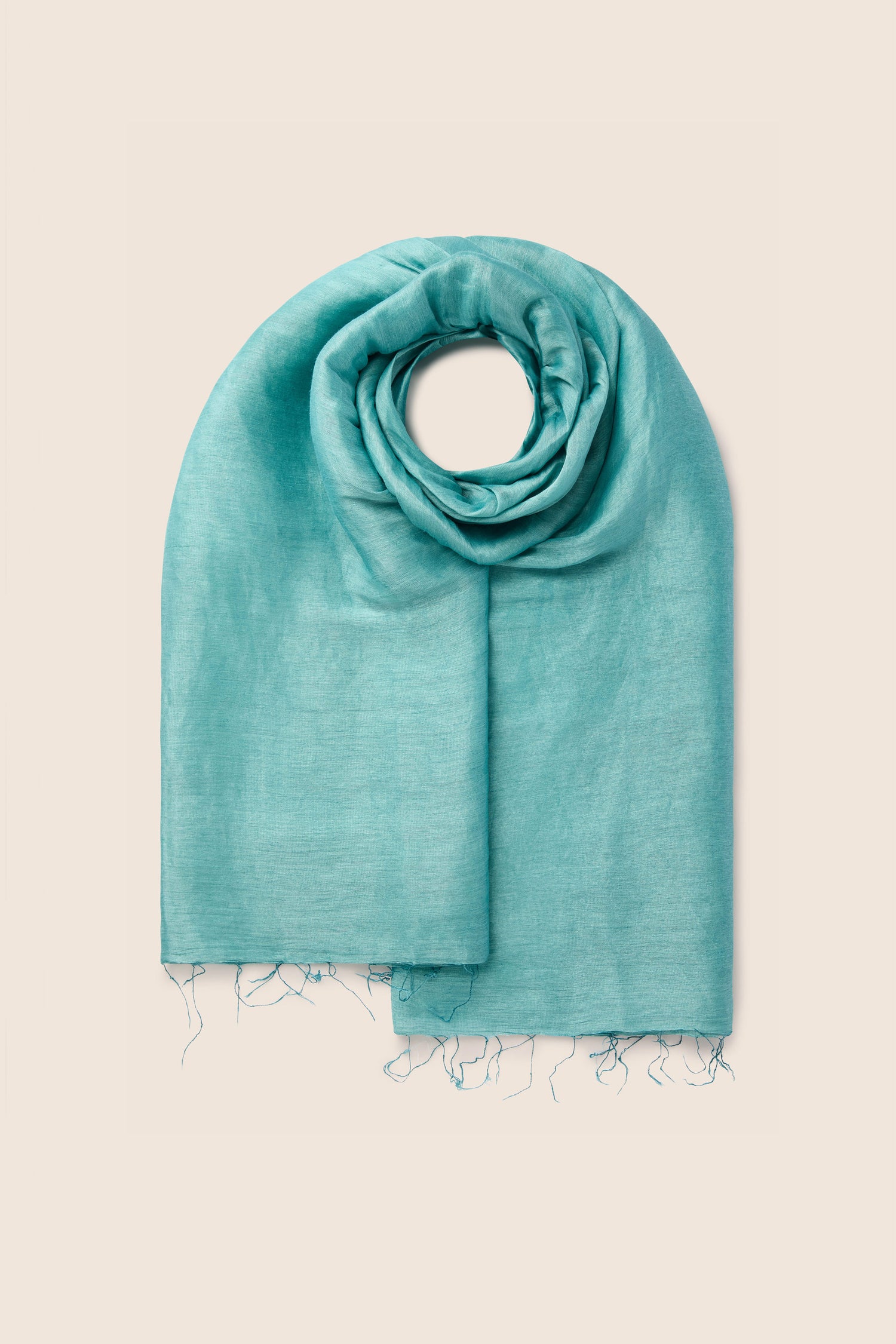 A Soft Linen Scarf in a folded teal hue with frayed edges rests elegantly on a light beige background, highlighting its luxurious linen quality and commitment to fair trade origins.