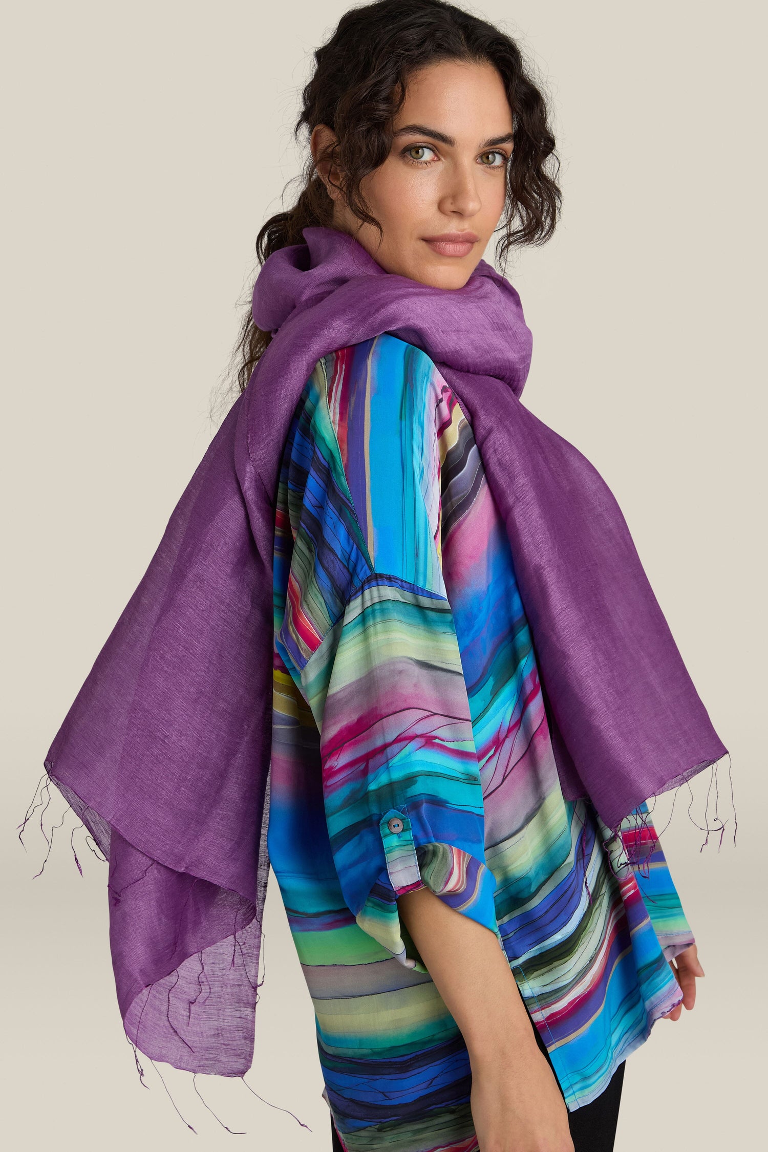 A woman with dark curly hair, wearing a vibrant multi-colored shirt, elegantly models the luxurious Soft Linen Scarf in purple as she gazes over her shoulder.