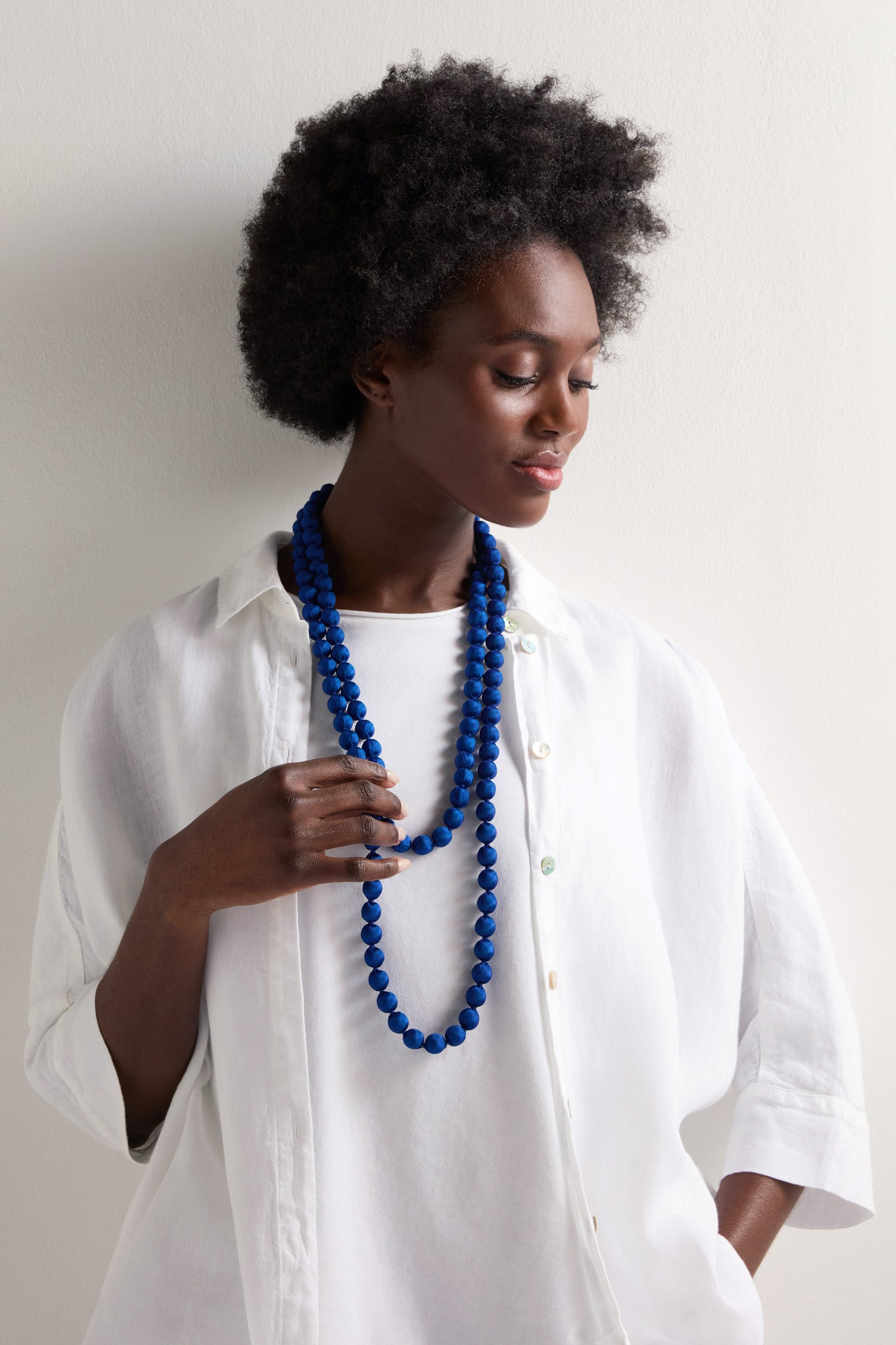 A person in a white shirt holds the Handmade Long Silk Spheres Necklace, showcasing its vibrant blue beads and artisanal craftsmanship while gazing downward.