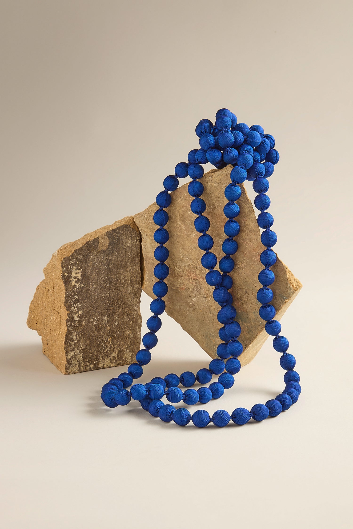 The Handmade Long Silk Spheres Necklace, vibrant blue and expertly crafted, is elegantly draped over two pieces of broken stone against a plain background.