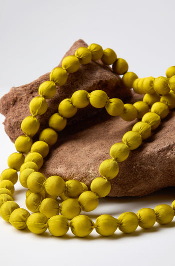 A Handmade Long Silk Spheres Necklace, displaying vibrant colors, is elegantly arranged over two uniquely shaped brown stones on a simple white backdrop.