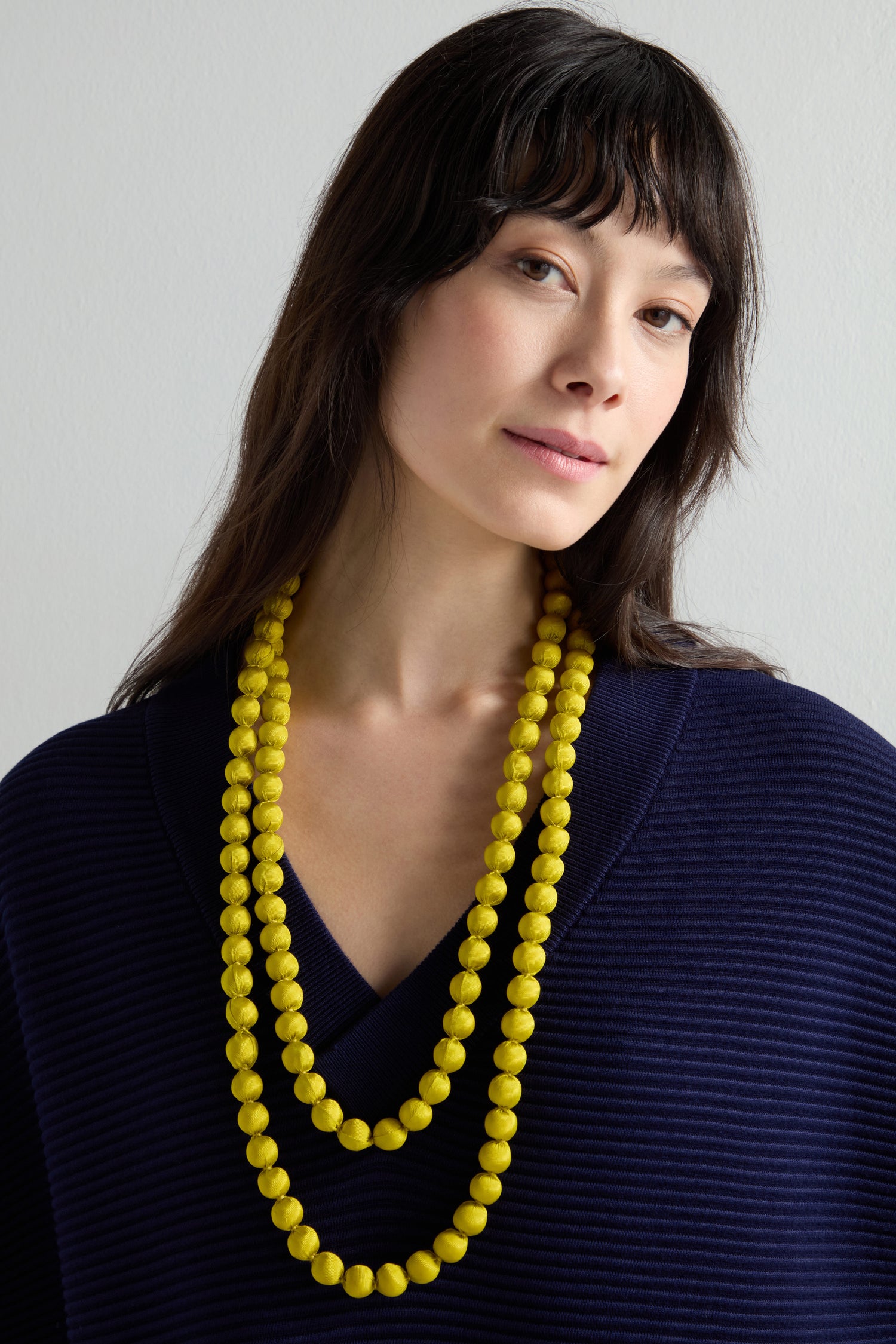 A person stands against a plain background wearing a navy top and a handmade long necklace called the "Handmade Long Silk Spheres Necklace" with yellow silk spheres.