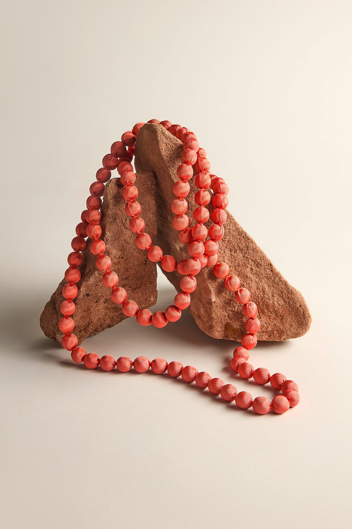 The Handmade Long Silk Spheres Necklace, in coral, drapes elegantly over two triangular rock formations on a neutral background, showcasing artisanal craftsmanship.