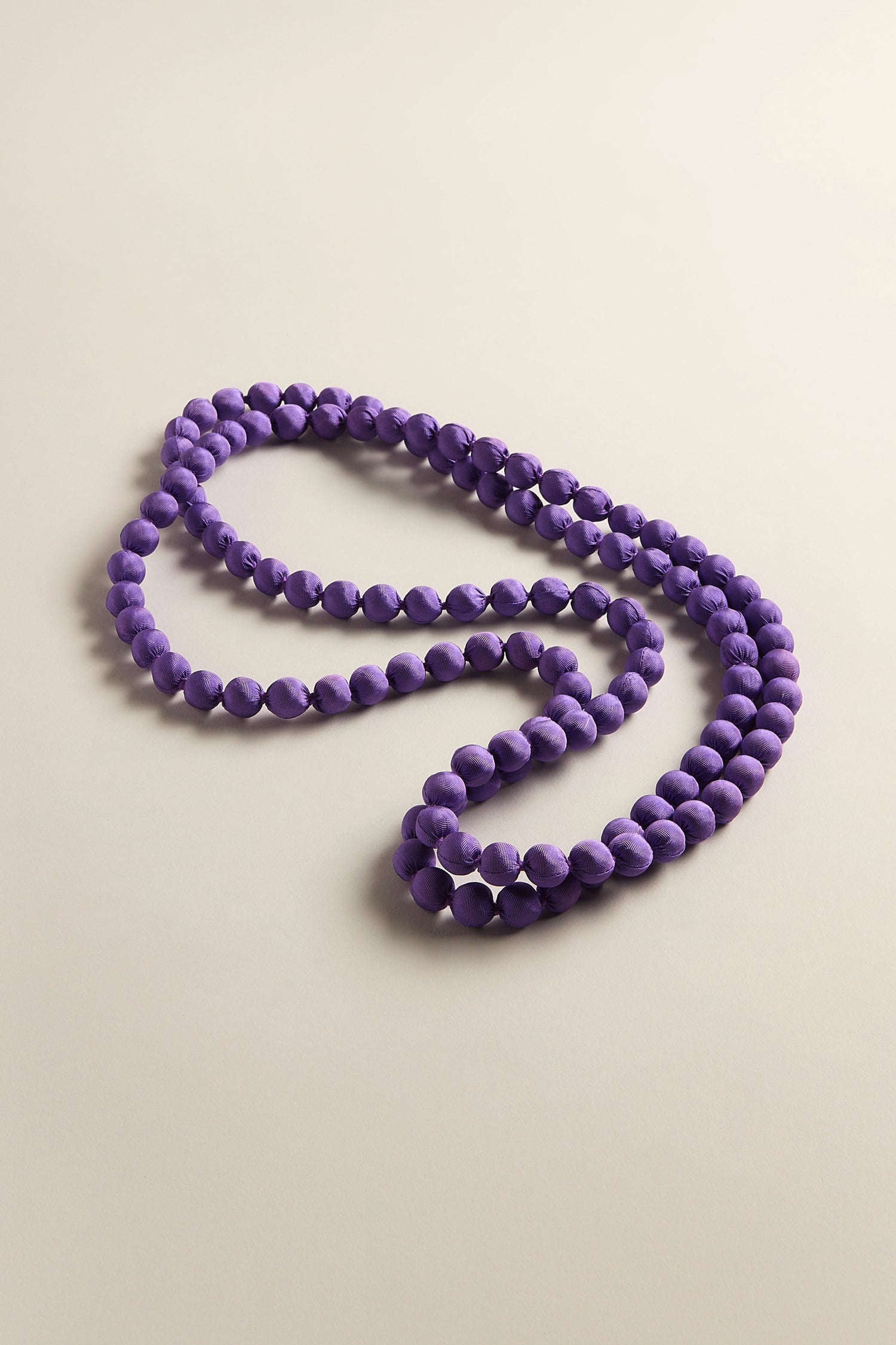 The Handmade Long Silk Spheres Necklace, with its purple bead strand, spirals loosely on a light surface, highlighting vibrant craftsmanship and unique accessories.