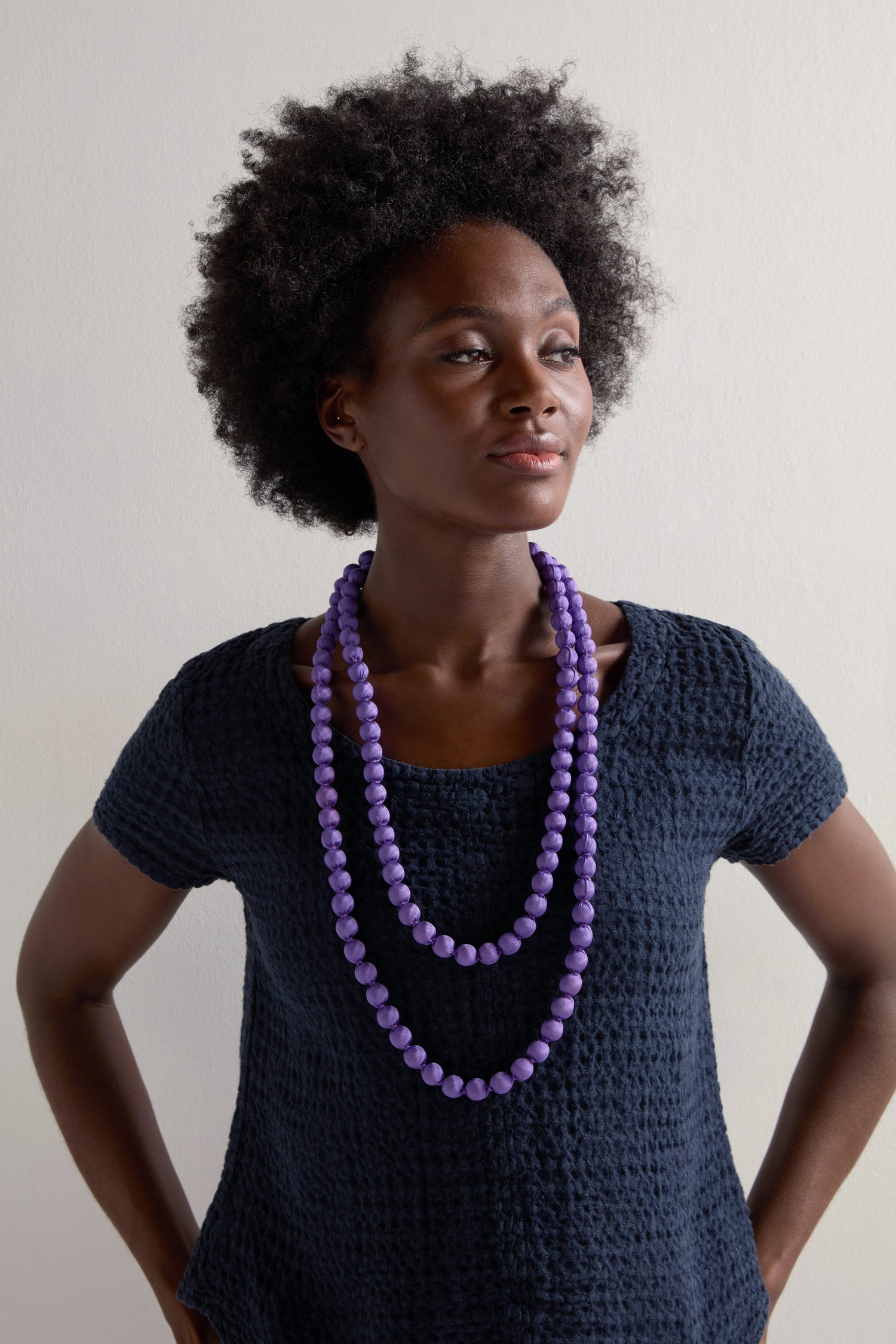 A person wearing a textured dark shirt and two strands of purple beads showcases vibrant accessories, including a Handmade Long Silk Spheres Necklace, against a plain background.