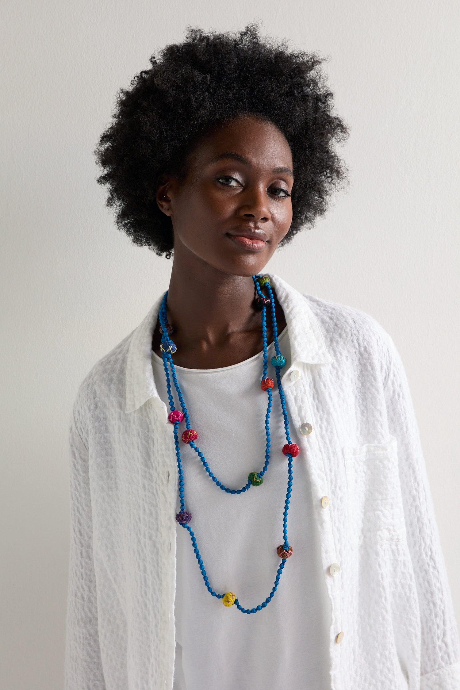 A person wearing a white textured shirt and the Handmade Rainbow Silk Spheres Necklace, showcasing vibrant colorful beads, stands in front of a plain white background.