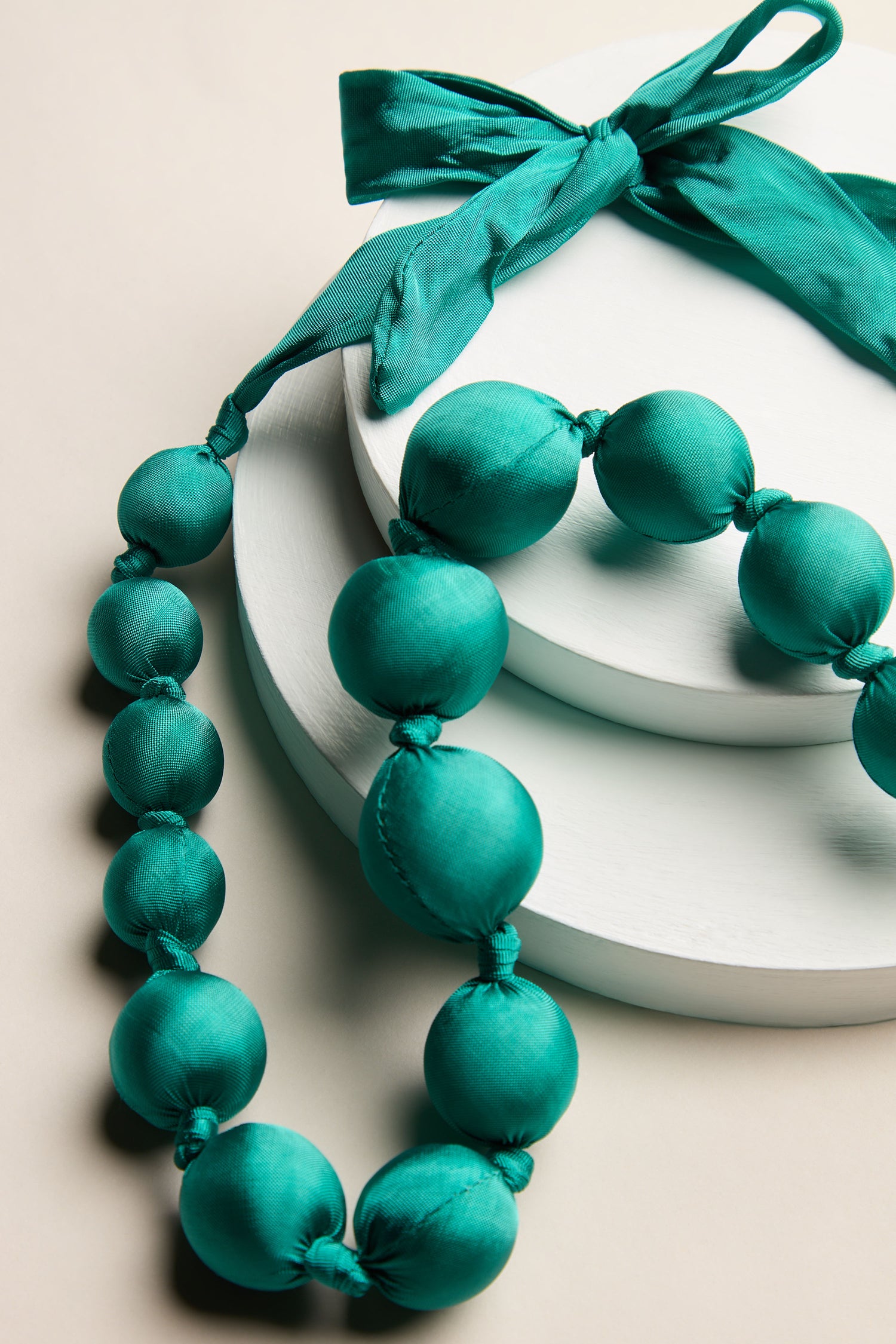 A Handmade Silk Spheres Necklace in a stunning teal hue, adorned with matching bows and showcasing artisanal craftsmanship, is elegantly displayed on two round white stands.