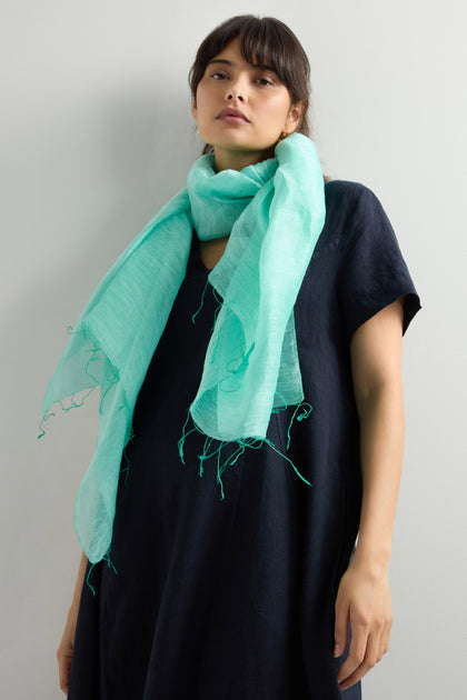 A person elegantly wearing a Spearmint Soft Linen Fringe Scarf and a black dress stands against a plain background, effortlessly showcasing the versatility of this stylish accessory.