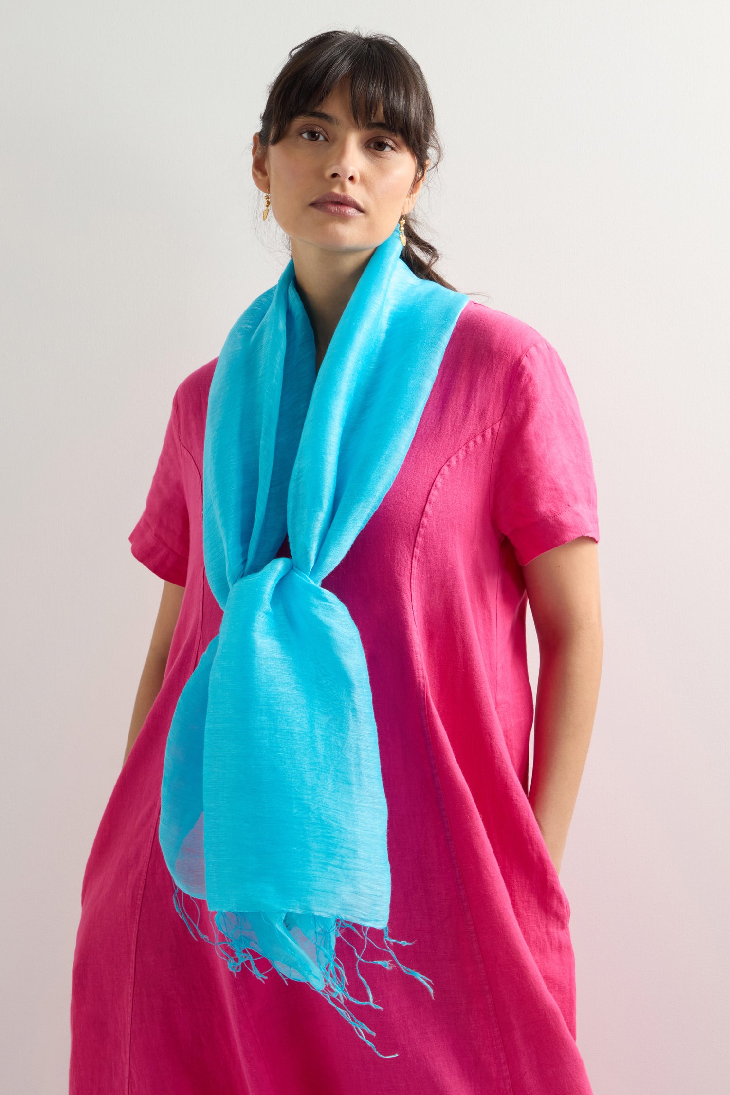 Against a plain background, a person wears a pink dress with pockets and accessorizes with the Soft Linen Fringe Scarf, infusing their versatile wardrobe with bohemian flair.