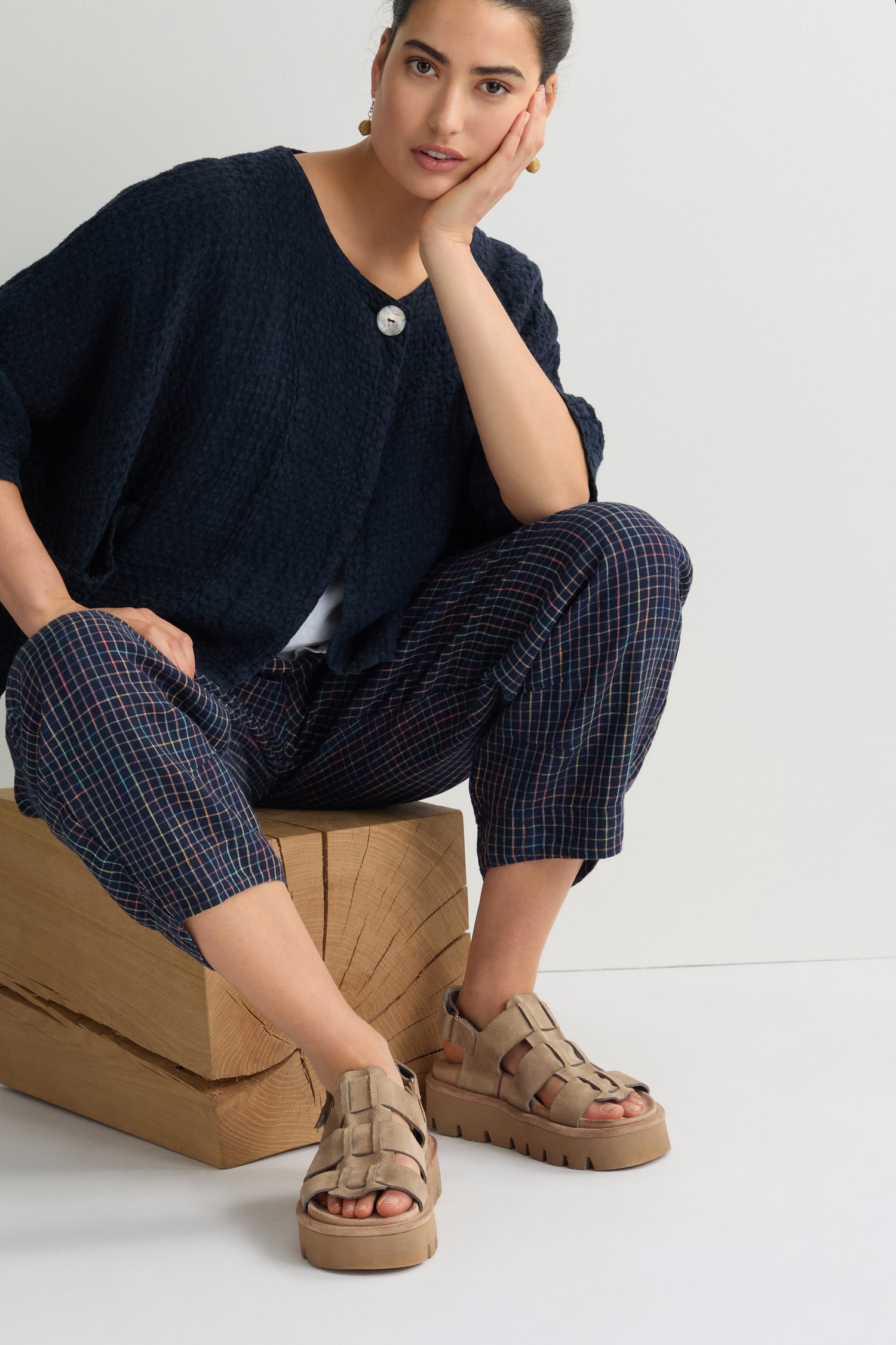 A person sits on wooden blocks, wearing a stylish black sweater and plaid pants, perfectly complemented by beige Suede Platform Velcro Sandals.