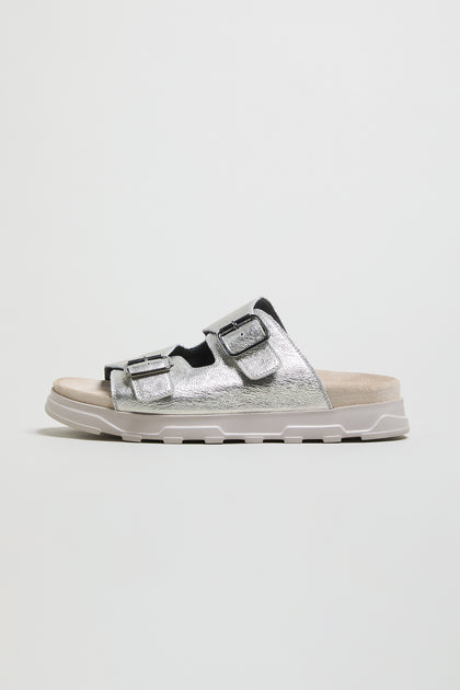 The Double Buckle Leather Sliders in metallic silver feature two adjustable straps and a cushioned sole, shown on a plain white background.