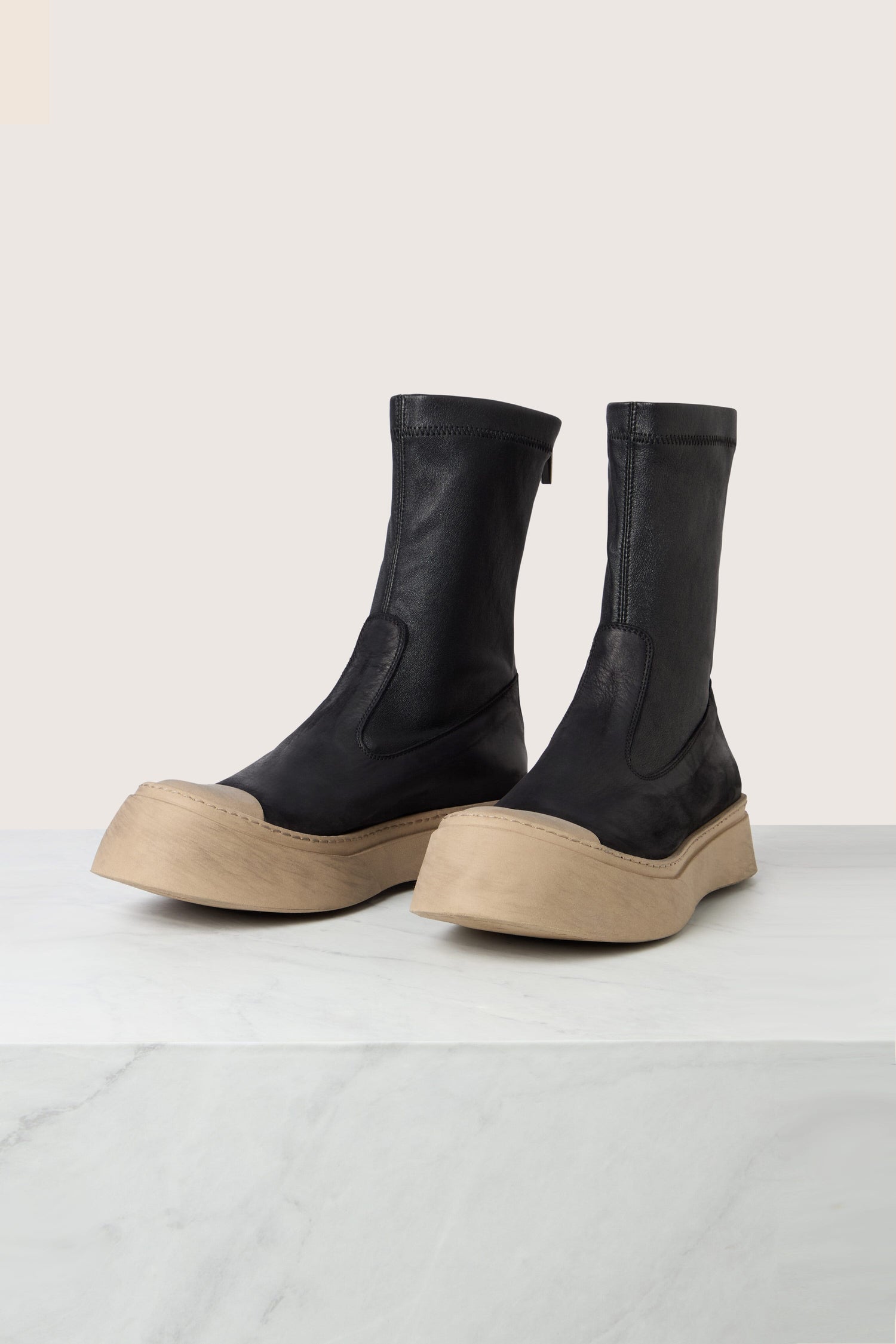 A pair of black Contrast Leather Stretch Boots, crafted from ultra-soft calfskin and featuring thick beige soles, are displayed on a white marble surface against a light background.